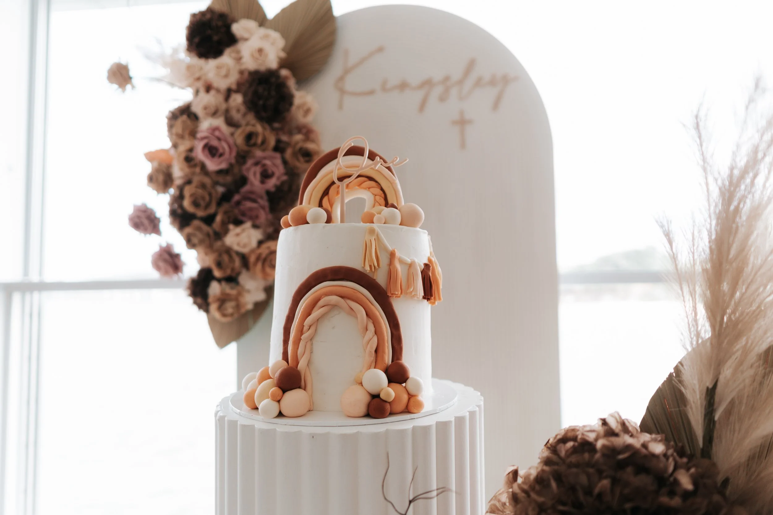 A two-tiered white cake decorated with rainbows and balls, with flowers and foliage in the background, set against a white backdrop with the word 'Gather' and a cross written on it.