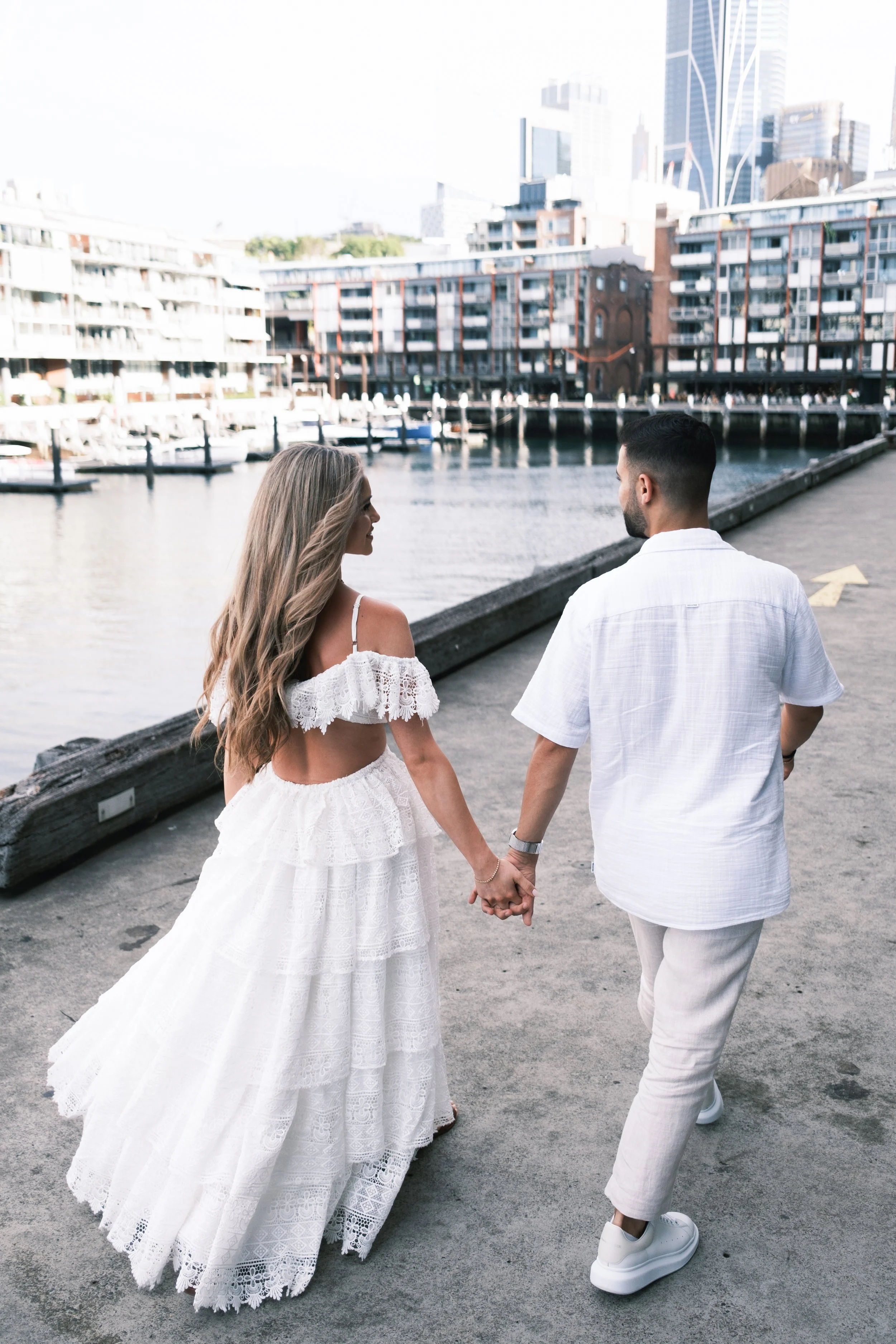 A couple walking hand in hand by a waterfront, with city buildings in the background, during daytime.