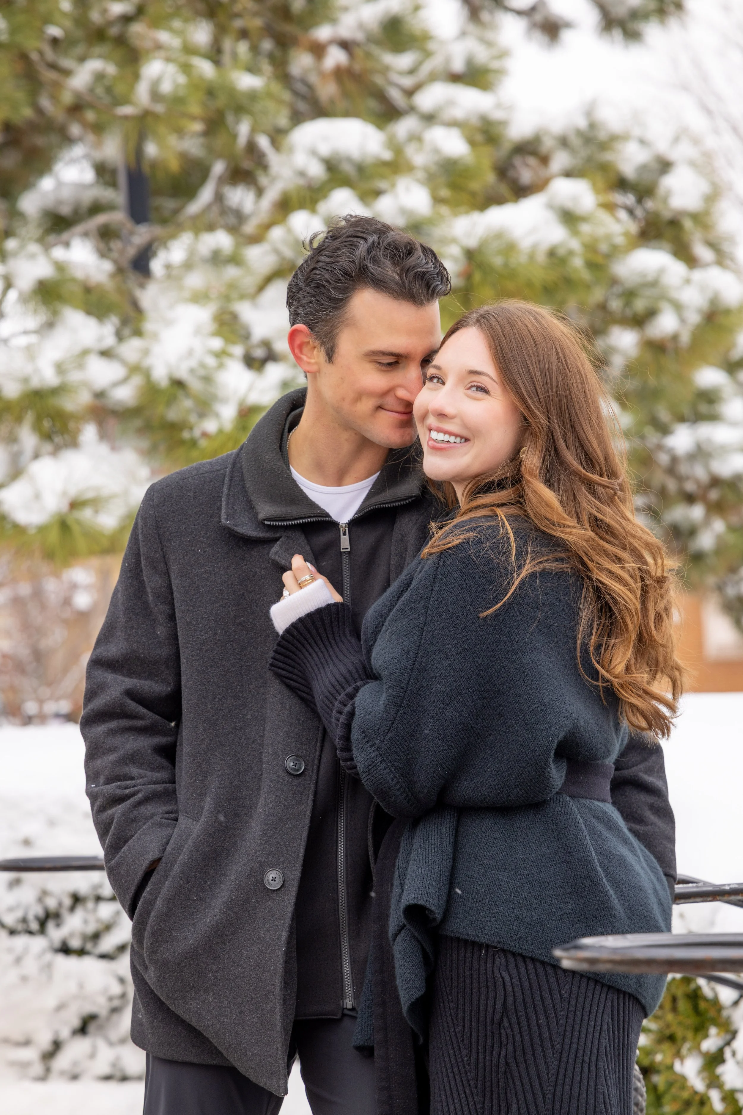 A young couple with light skin and brown and black hair standing closely together outdoors in a snowy park, smiling and embracing, with snow-covered trees in the background.