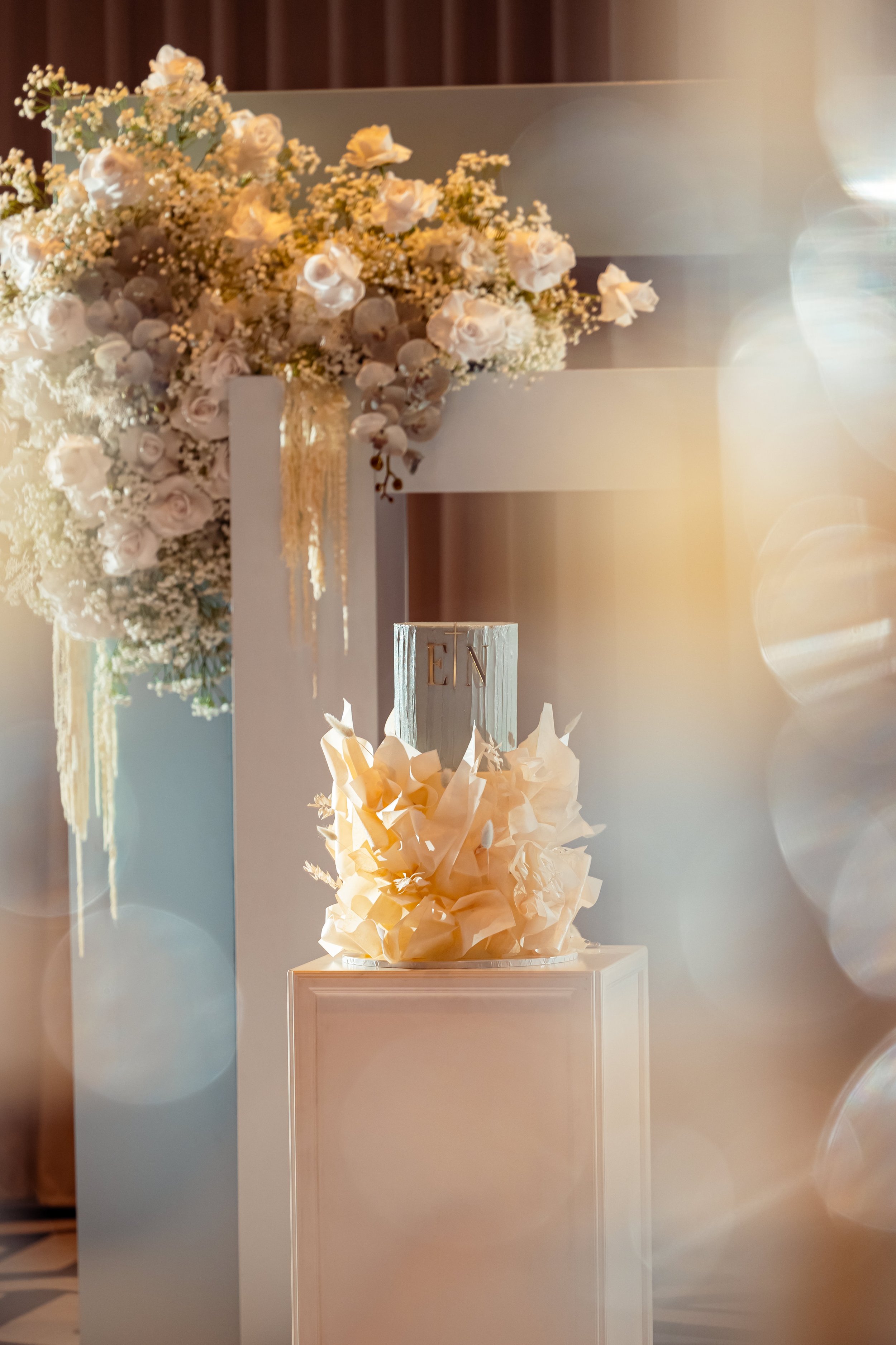 Wedding or event decor featuring a large flower arrangement of white roses and baby's breath, and a candle on a pedestal with the initials "TEN" in gold, set against a softly lit background with circles of light.