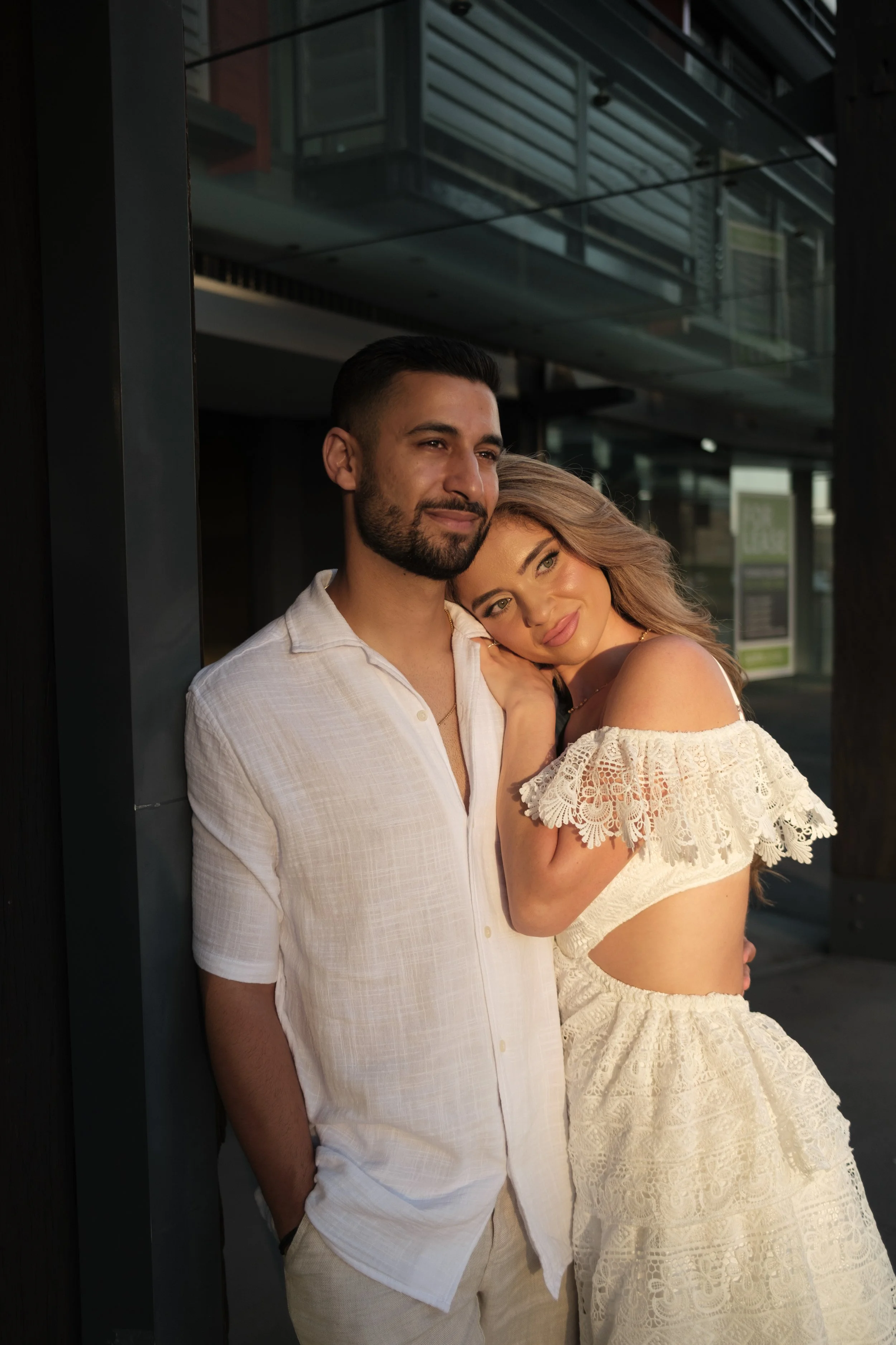 A man and woman standing close together outdoors, with the woman's head resting on the man's shoulder, both smiling softly. The woman is wearing a white off-the-shoulder dress with lace details, and the man is in a light-colored short-sleeve shirt.