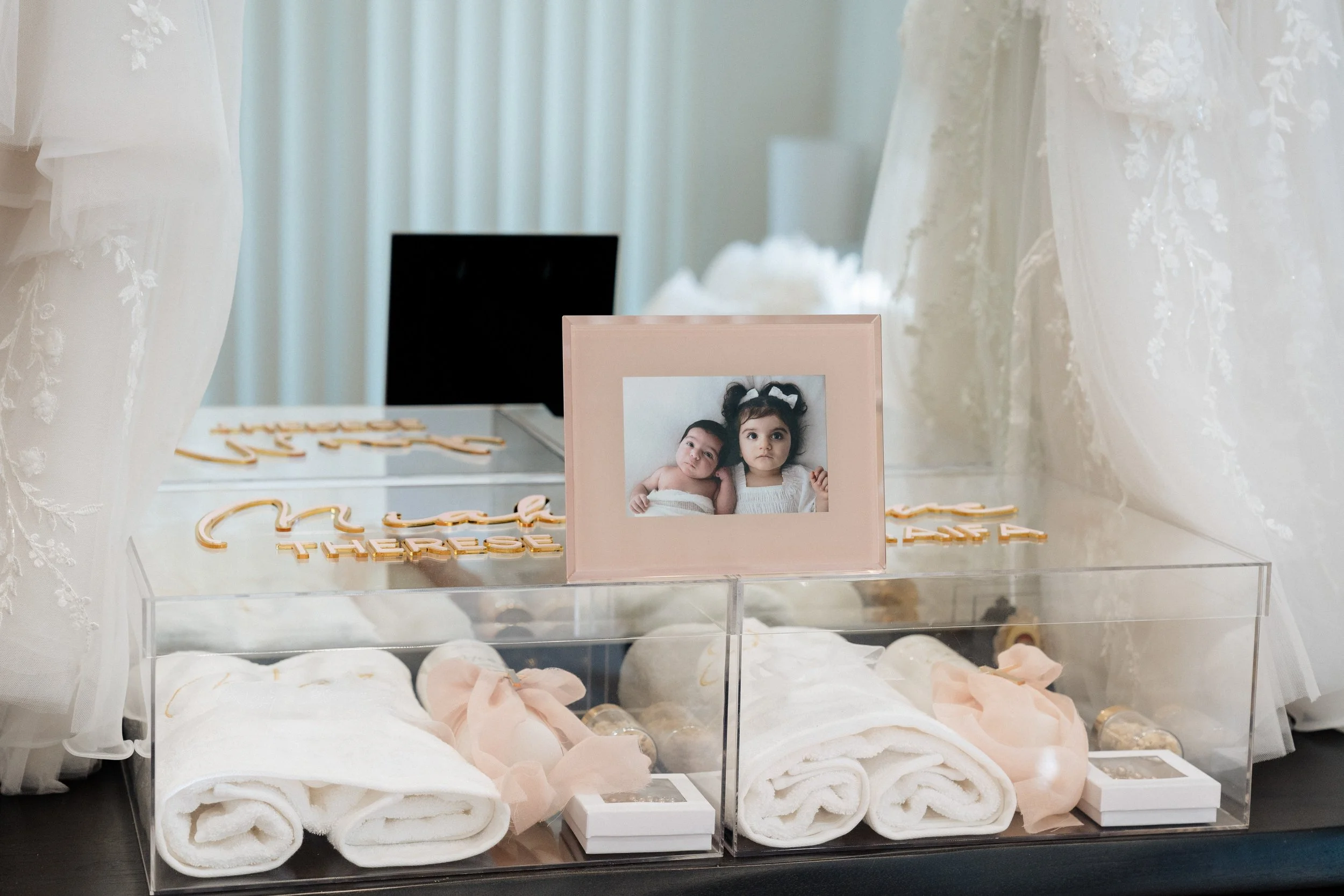 A clear acrylic box with white rolled towels inside, decorated with light pink bows and small bottles, placed on a dark surface. A pink picture frame with a photo of two young children is in front. Behind, there are soft white curtains and a black sc