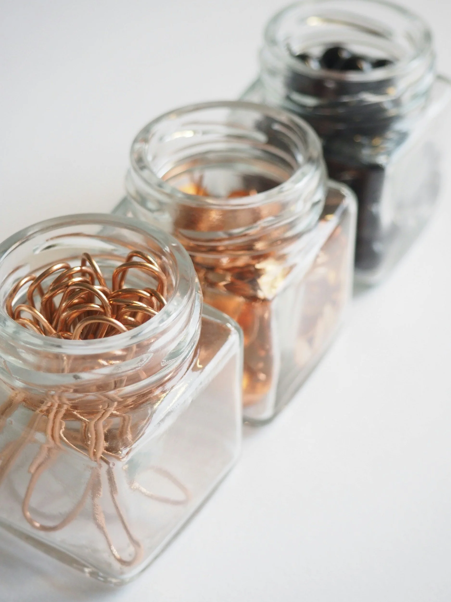 Three glass jars containing different types of metal paper clips, arranged in a row on a white surface.