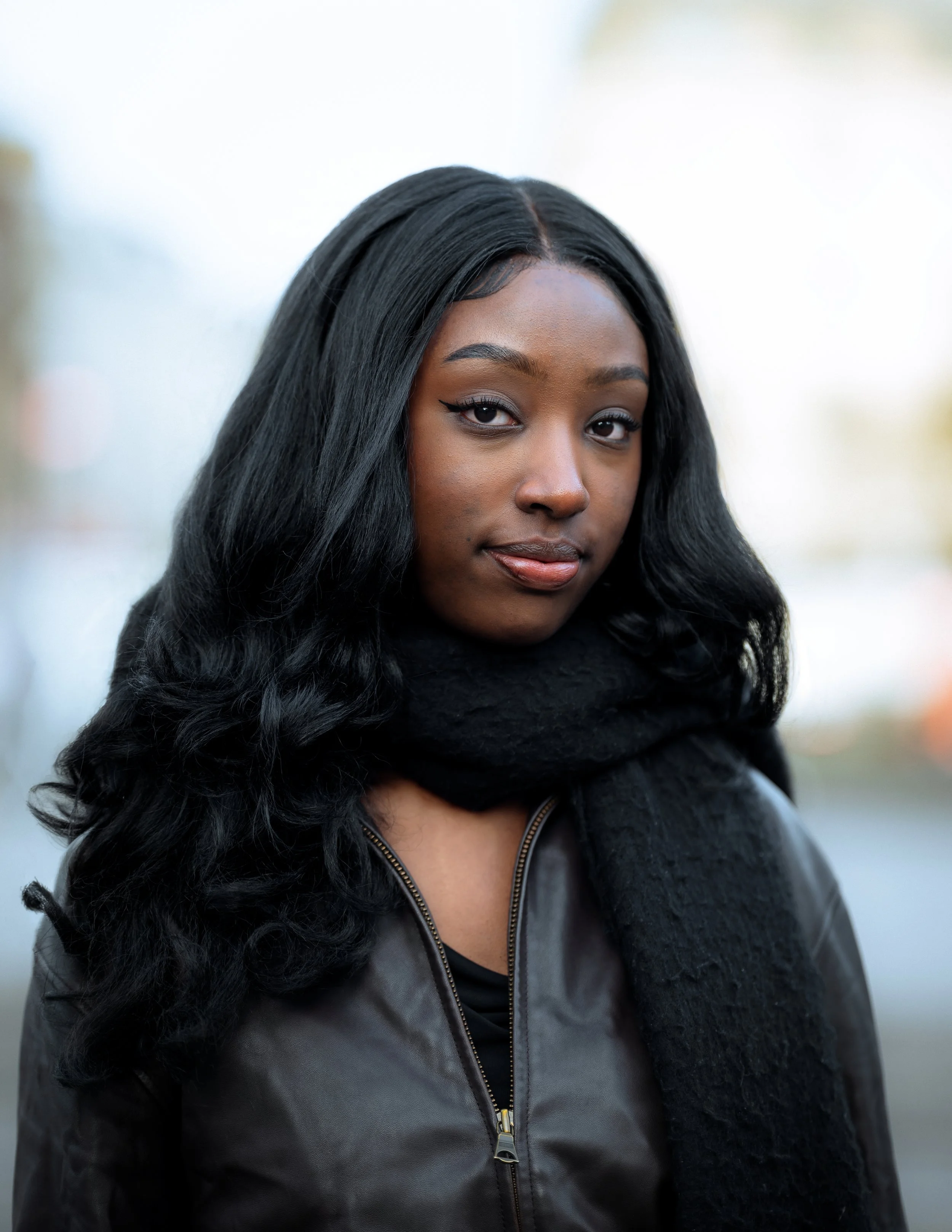 Portrait of a young woman with long black hair, wearing a black scarf and leather jacket, outdoors during daytime.