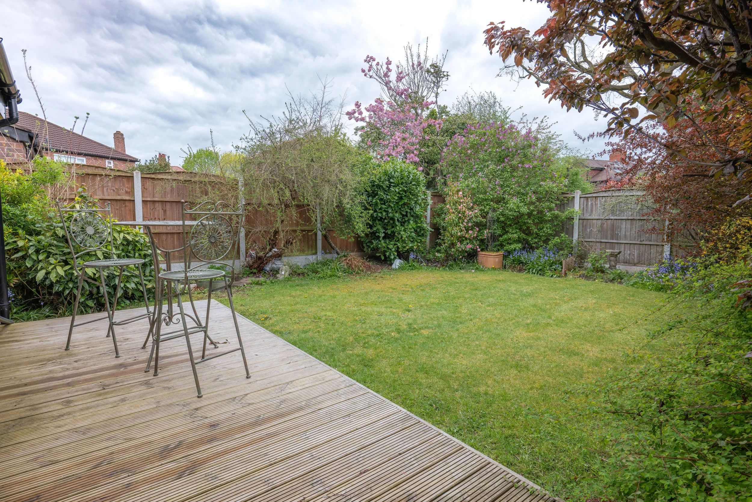 A backyard garden with a wooden deck and patio furniture, surrounded by trees and colorful flowering shrubs, enclosed by a wooden fence under a cloudy sky.