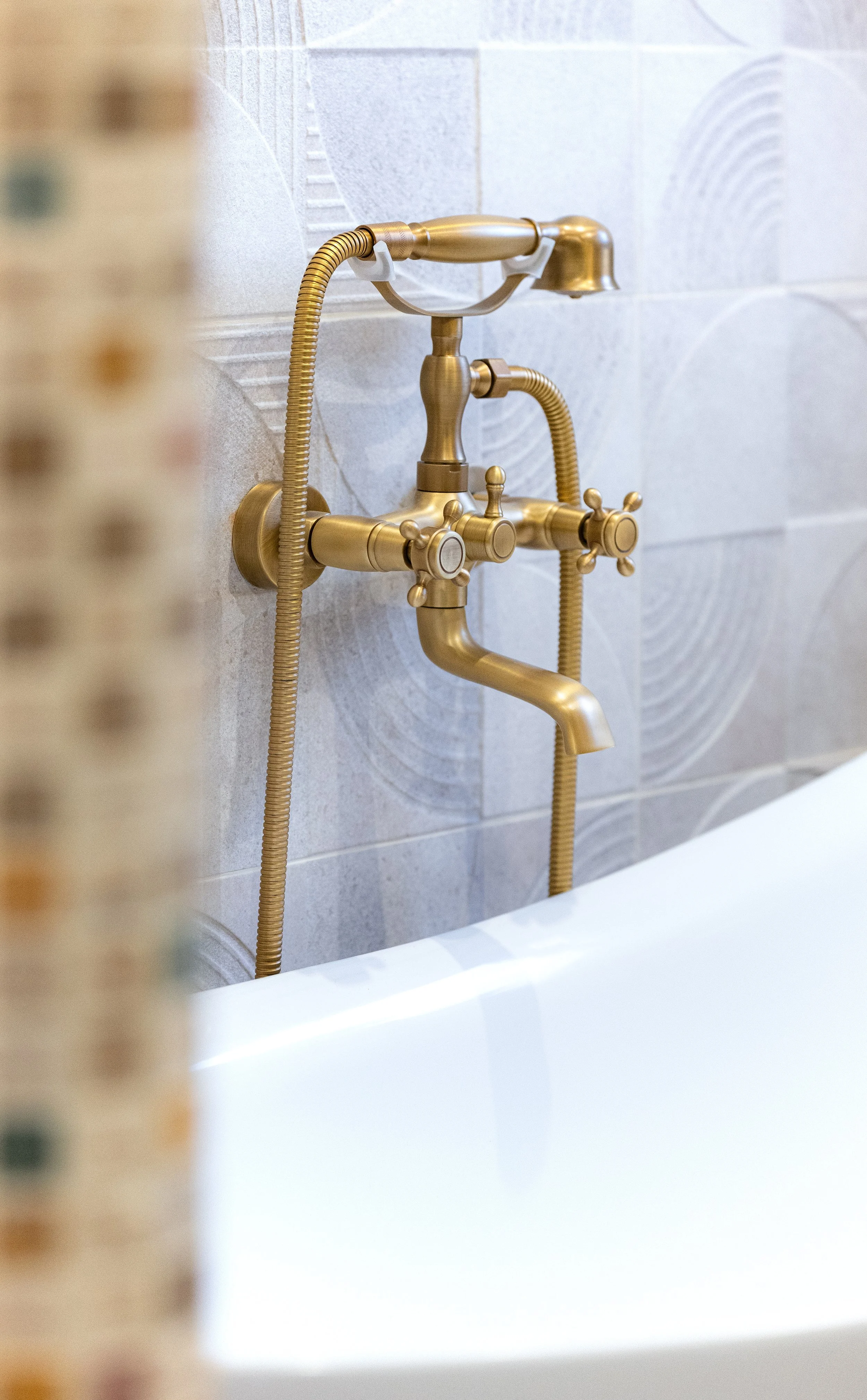 Brass bathtub faucet with dual handles and attached handheld showerhead mounted on a tiled bathroom wall.