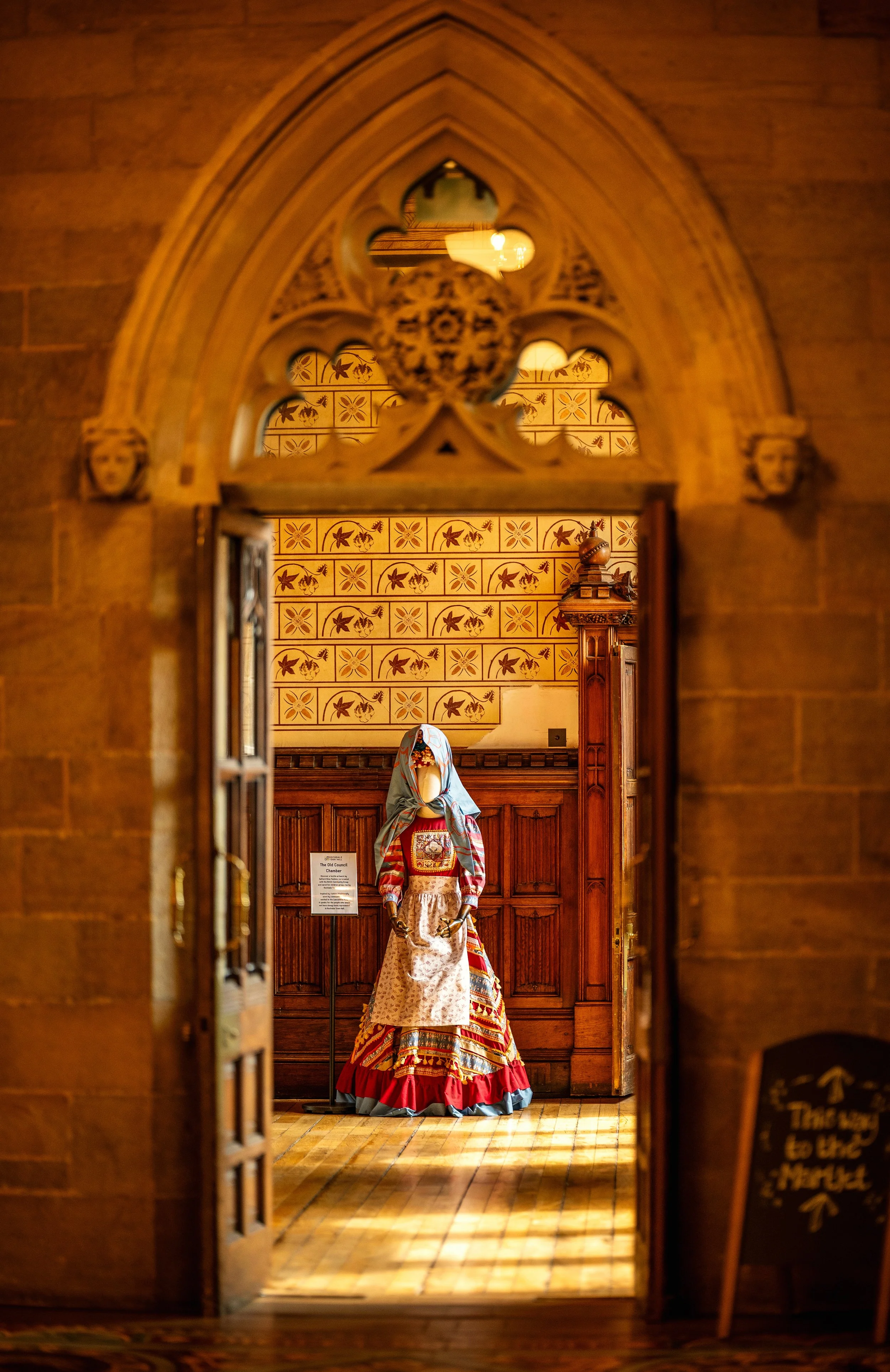 A mannequin dressed in traditional, colorful folk costume inside a wooden room, viewed through an ornate arched doorway.