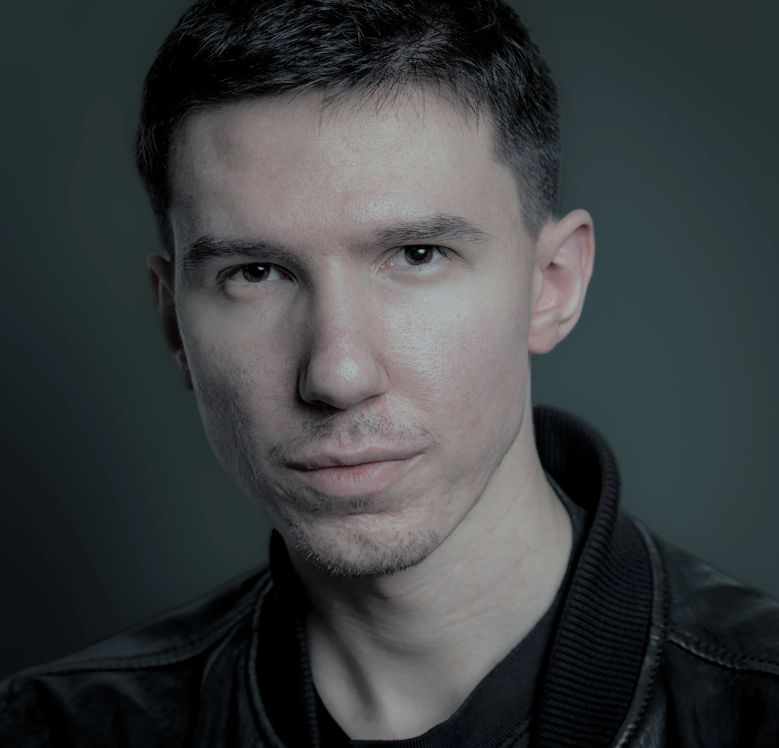 Close-up portrait of a young man with dark hair wearing a black leather jacket against a dark, neutral background.