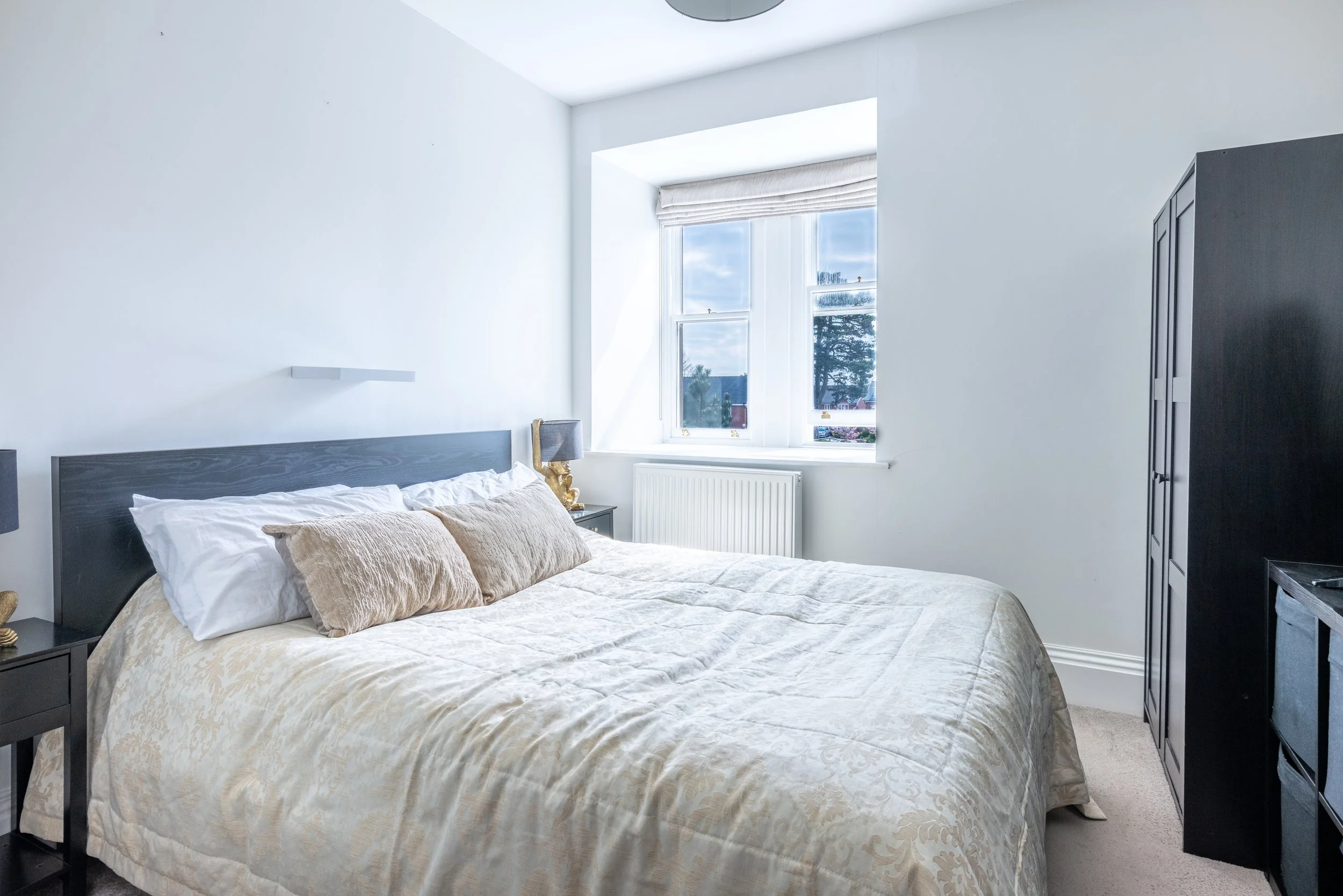 A bedroom with a large window, a bed with beige and white bedding, two beige pillows, black nightstands, and a black wardrobe.