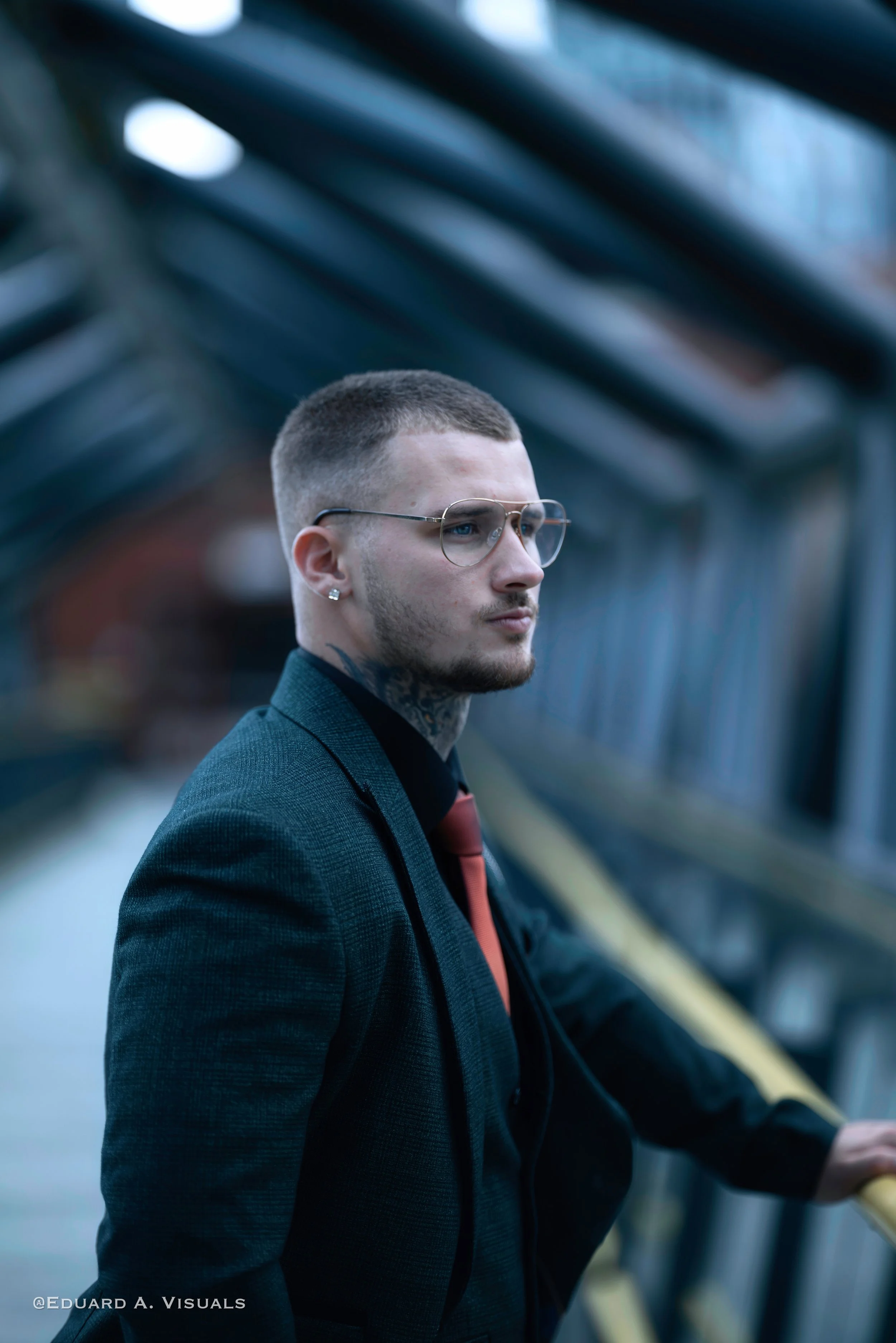 A man with short hair, glasses, and tattoos on his neck and ear, dressed in a dark suit with a tie, looking serious and standing on a bridge or walkway with glass and metal structure.