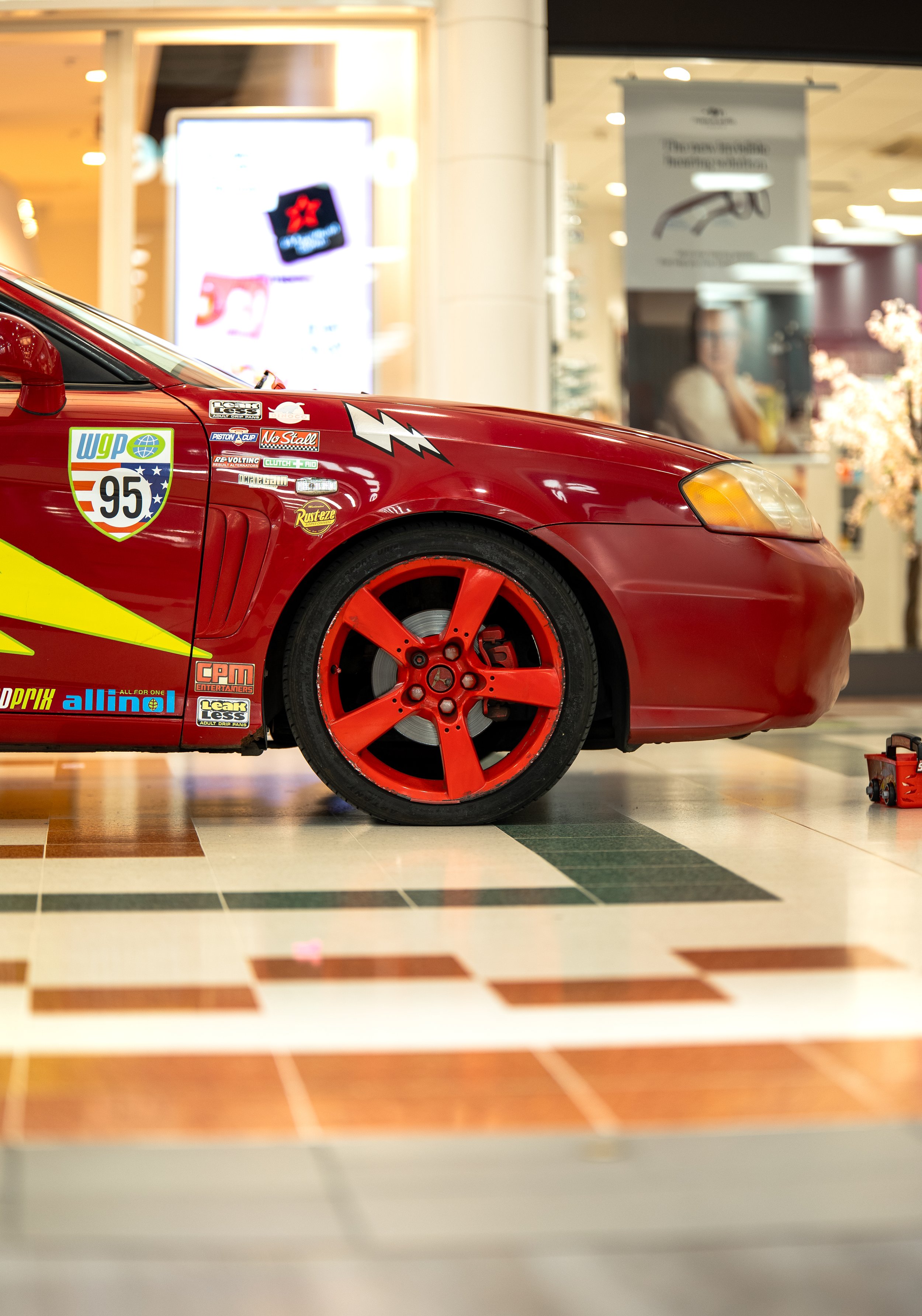 Indoor display of a red race car with red wheels and various sponsor stickers, parked on a checkered floor in a shopping mall.
