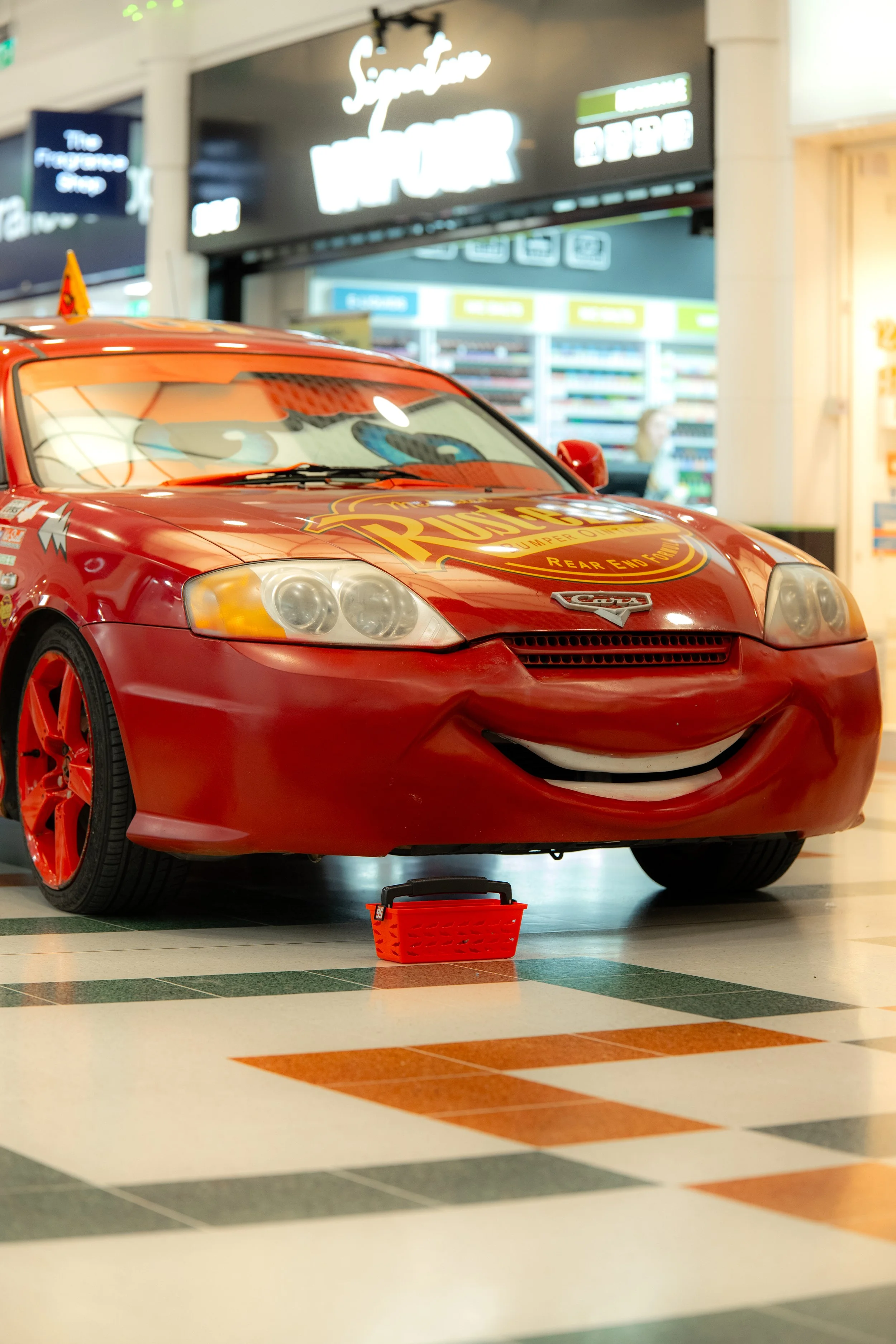 A red Lightning McQueen car from the movie Cars with smiling face and cartoon eyes, displayed indoors, with a small red shopping basket in front of it.