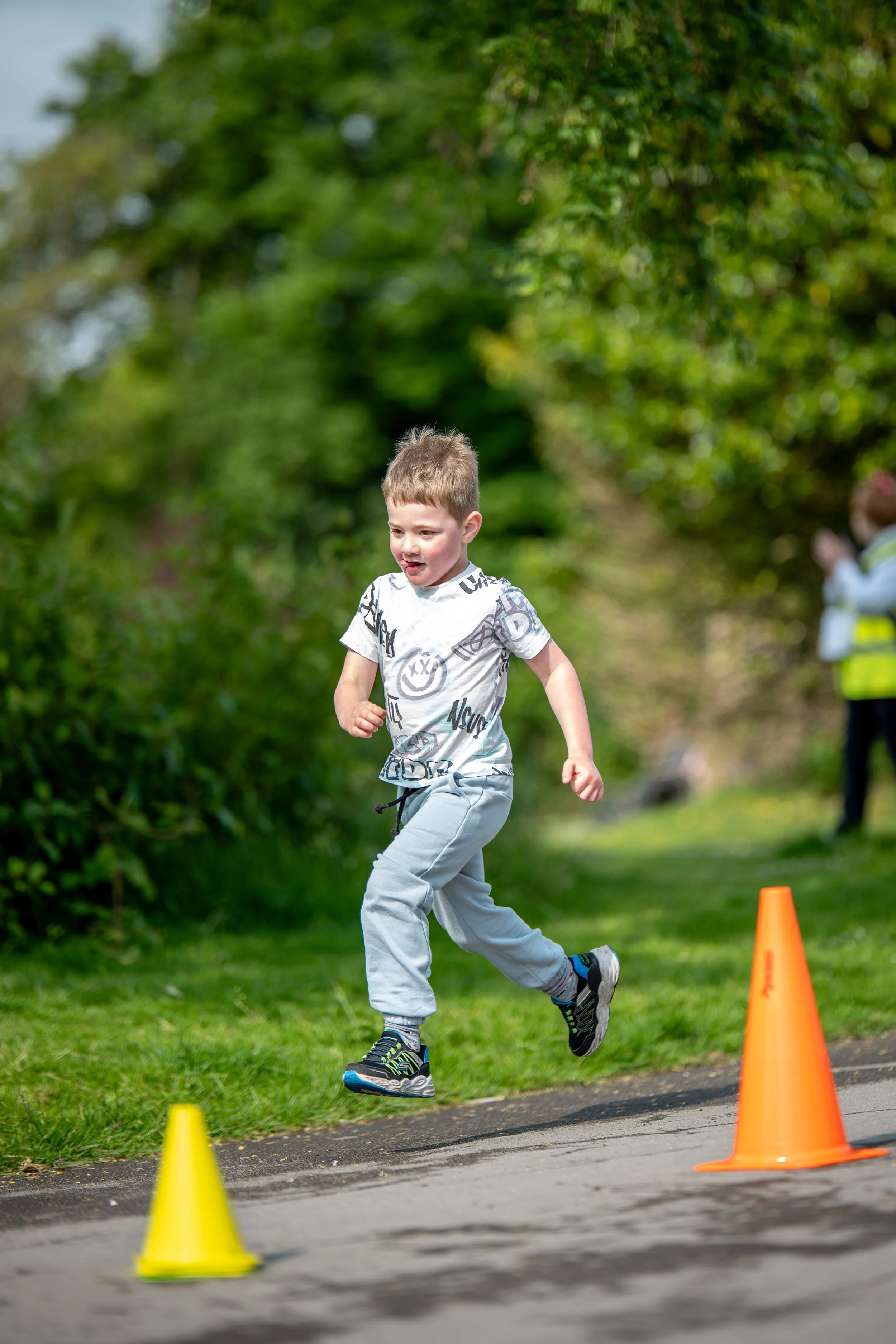 Young boy running outdoors on a path, participating in activity around orange and yellow cones, with greenery in the background.