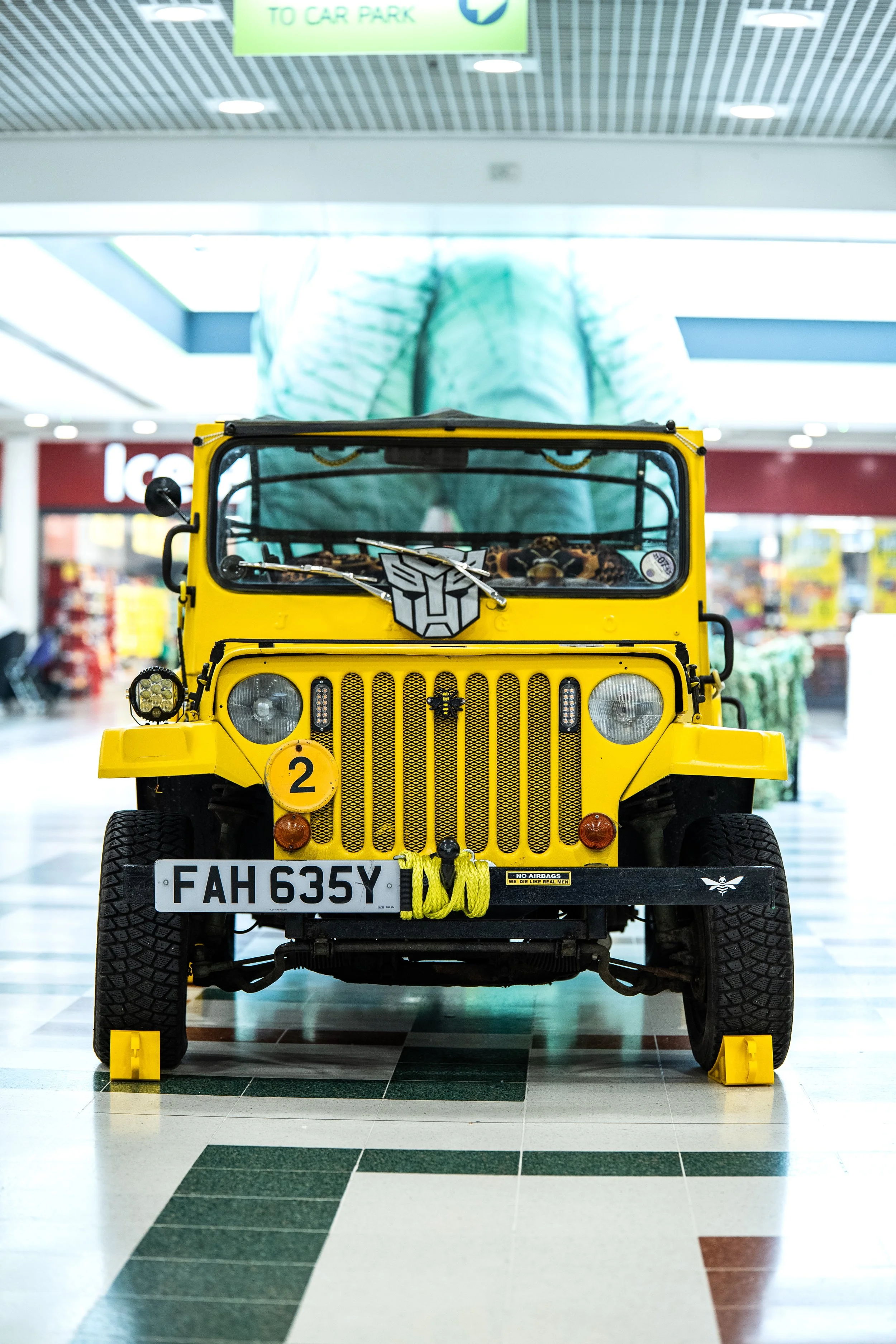 Front view of a yellow Jeep displayed indoors at a shopping mall with a large dinosaur statue in the background.