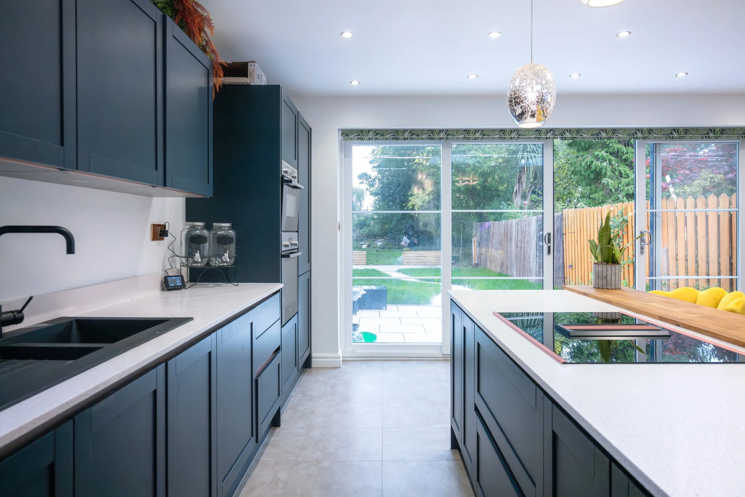 Modern kitchen with dark blue cabinets, white countertops, a sliding glass door leading to a backyard, and yellow bar stools at a wooden island.