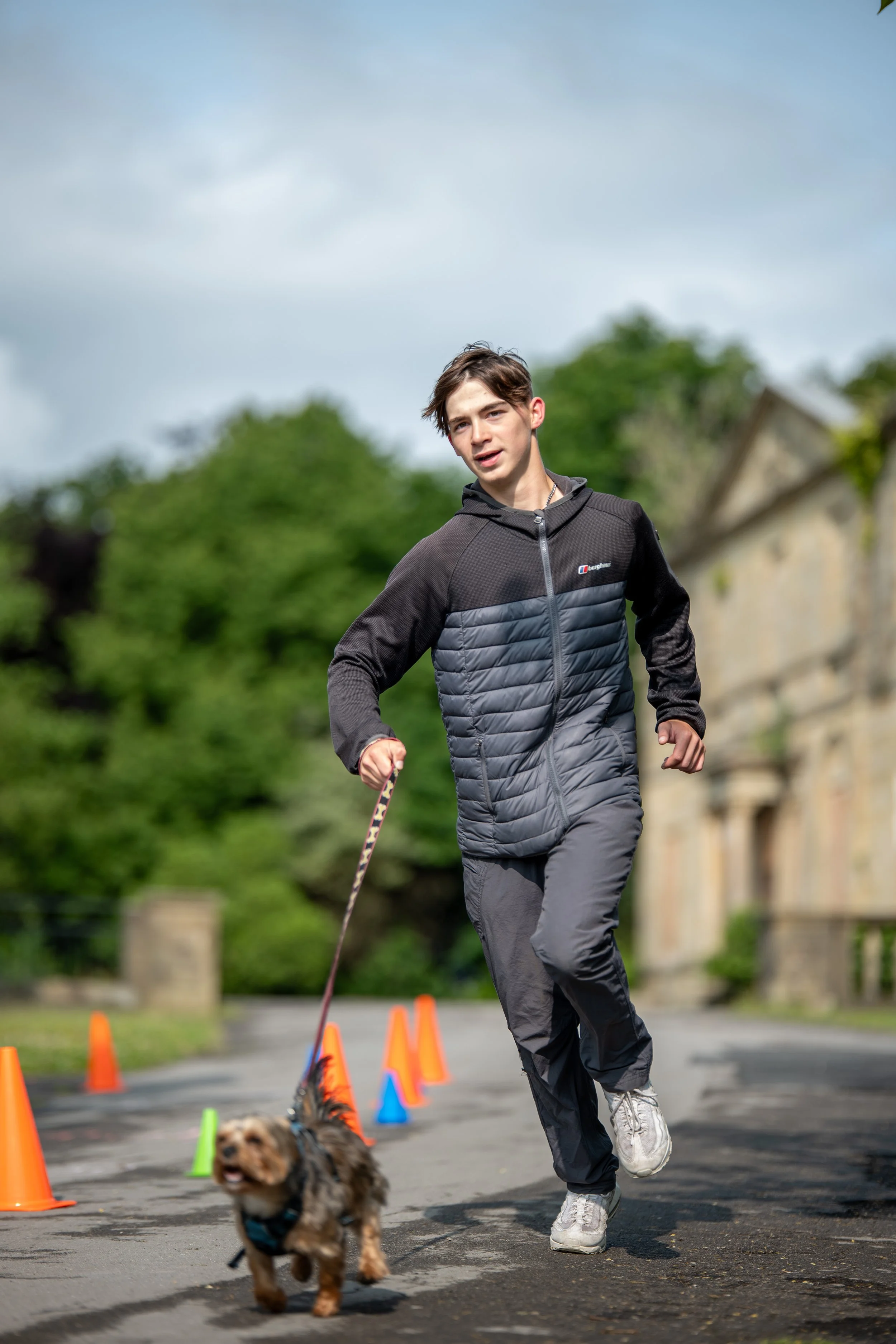 A young man in sportswear running outdoors with a small dog on a leash, surrounded by colorful traffic cones, on a paved path with greenery and a building in the background.