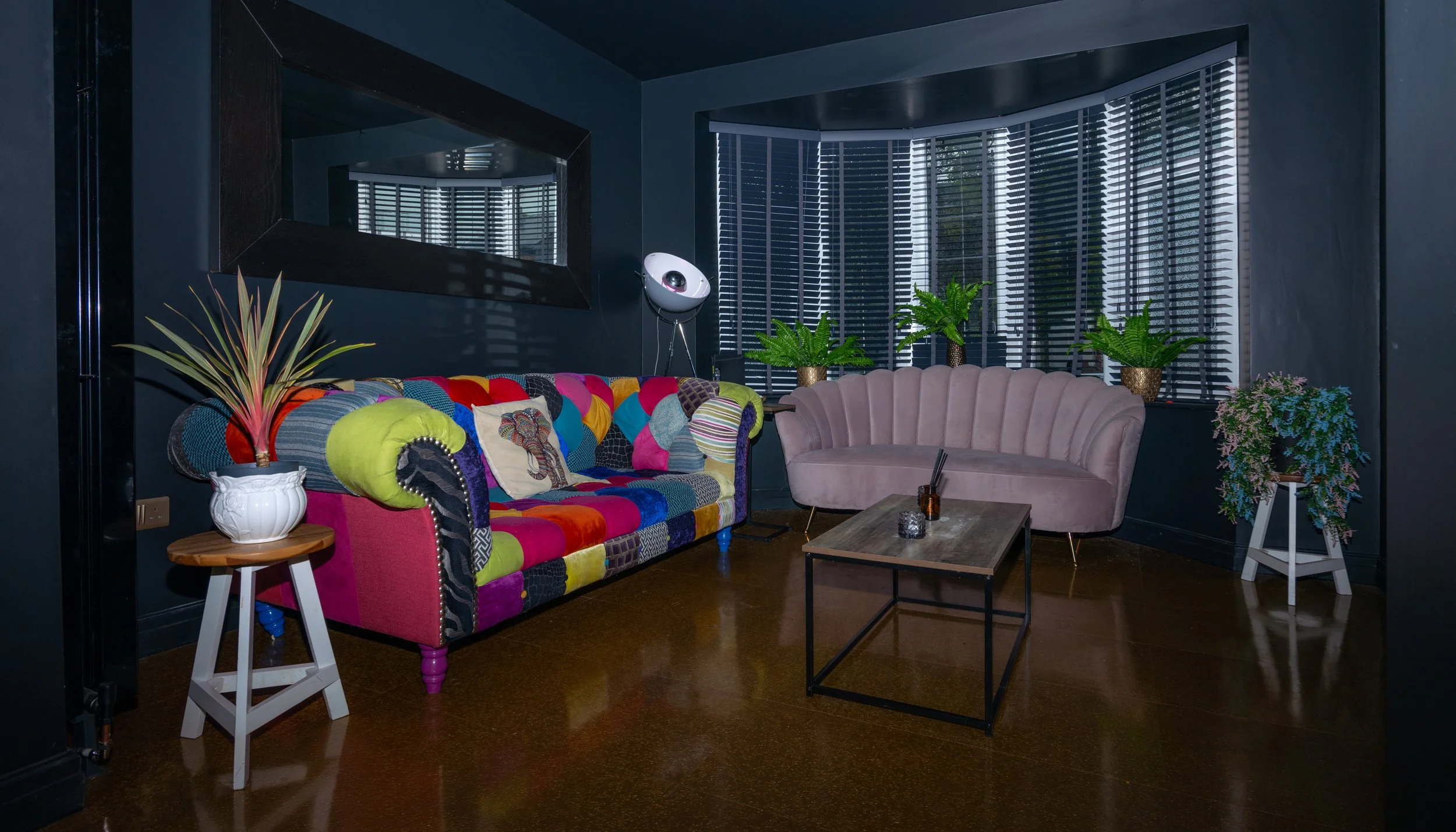 Colorful patchwork sofa, light pink velvet sofa, coffee table, potted plants, dark walls, window with blinds, mirror, floor lamp, wooden side table, decorative items.