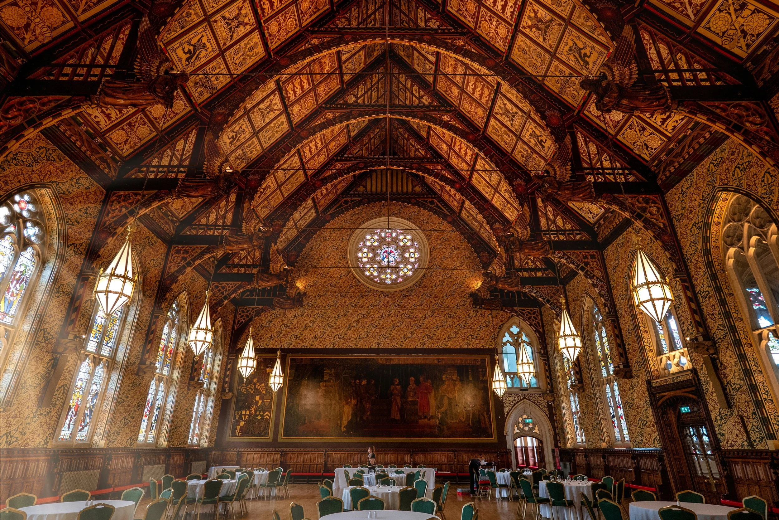 Interior of a grand, historic banquet hall with ornate, gilded woodwork and a high, arched ceiling featuring intricate patterns and a central stained glass window. The room is decorated with large paintings and has multiple round tables set for dinin