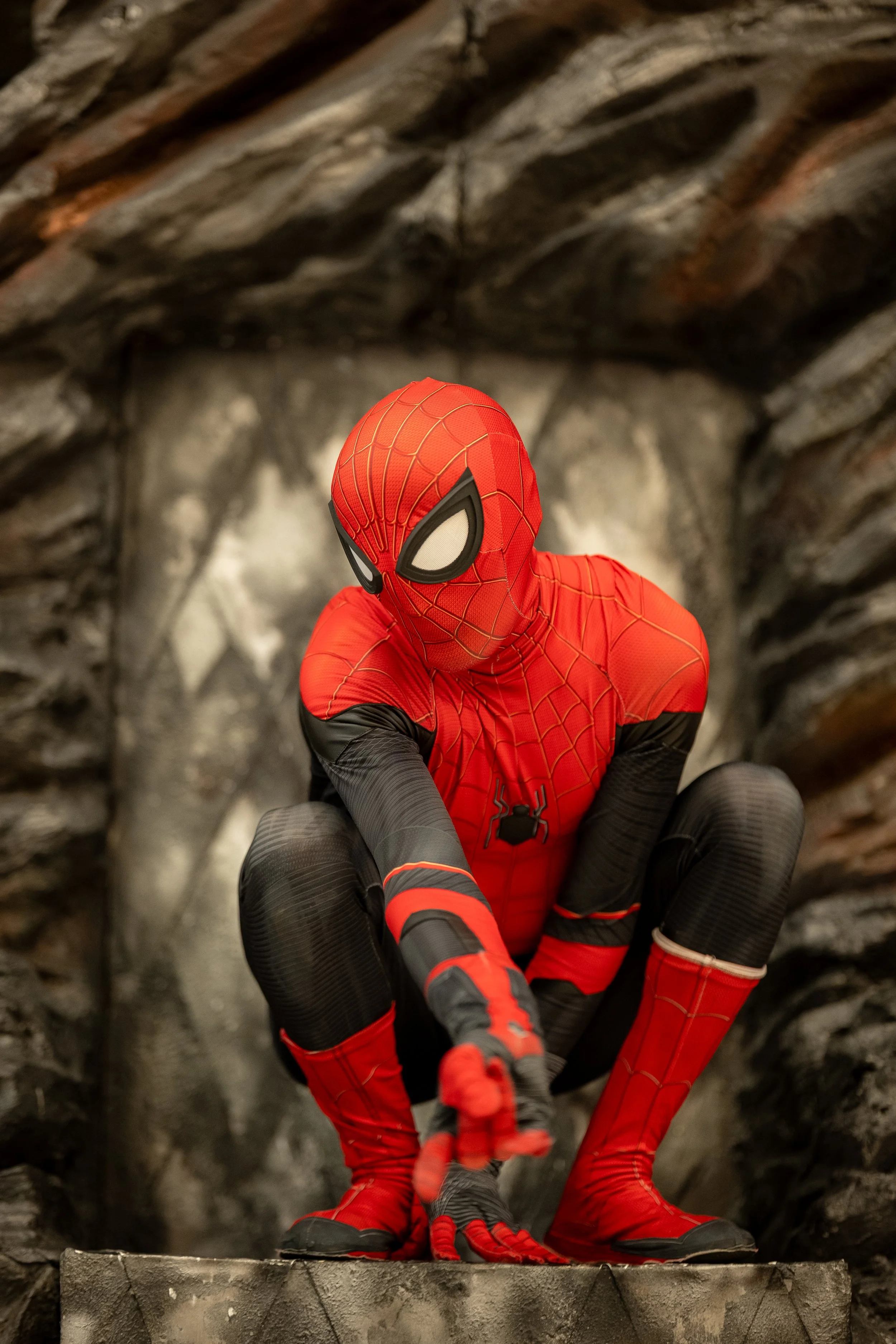 Person dressed as Spider-Man crouching on a stone surface, with a rocky or cave-like background.
