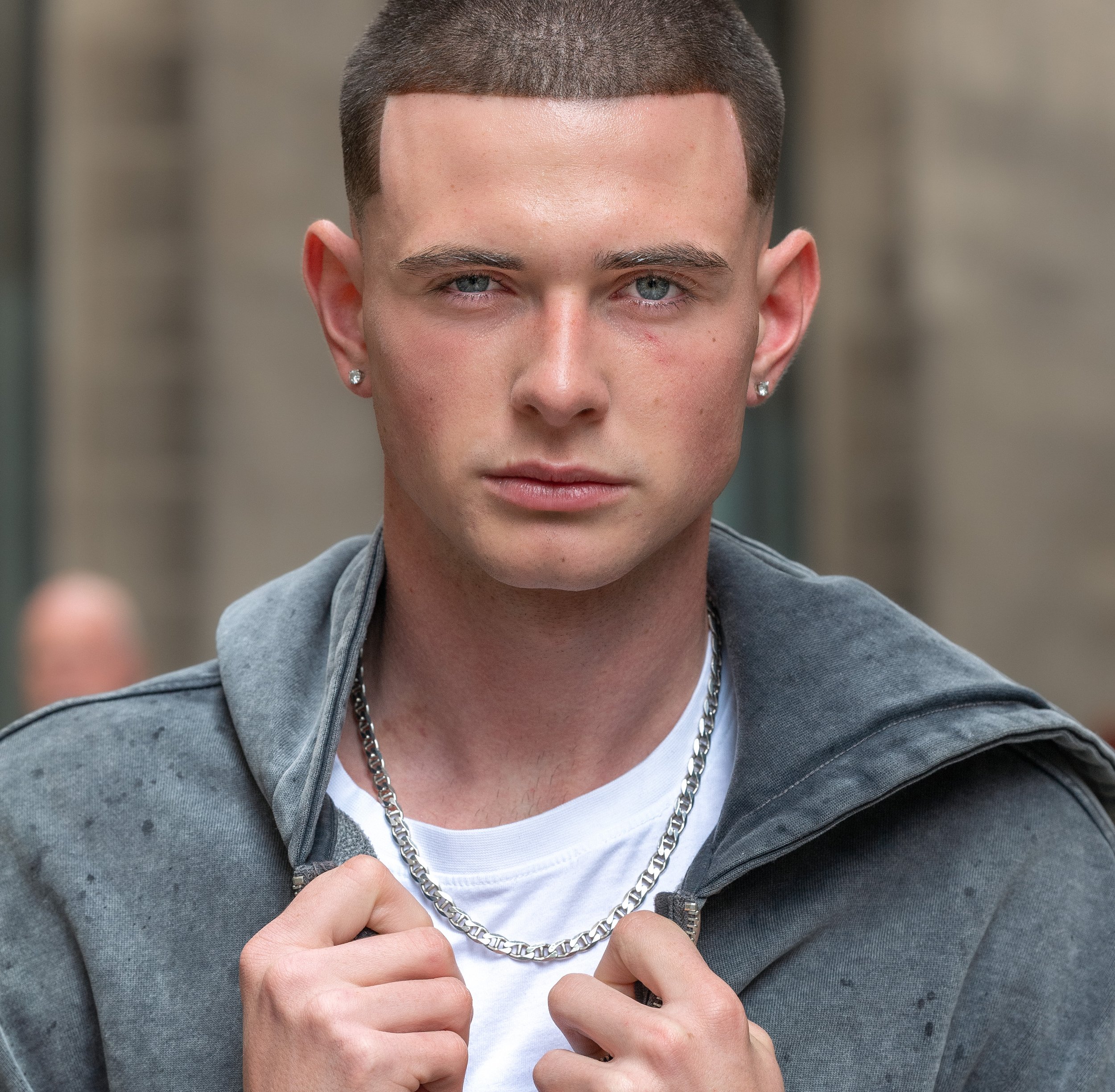 Close-up of a young man with short dark hair and light skin, wearing earrings, a chain necklace, a white T-shirt, and a gray jacket, pointing to his open zipper.