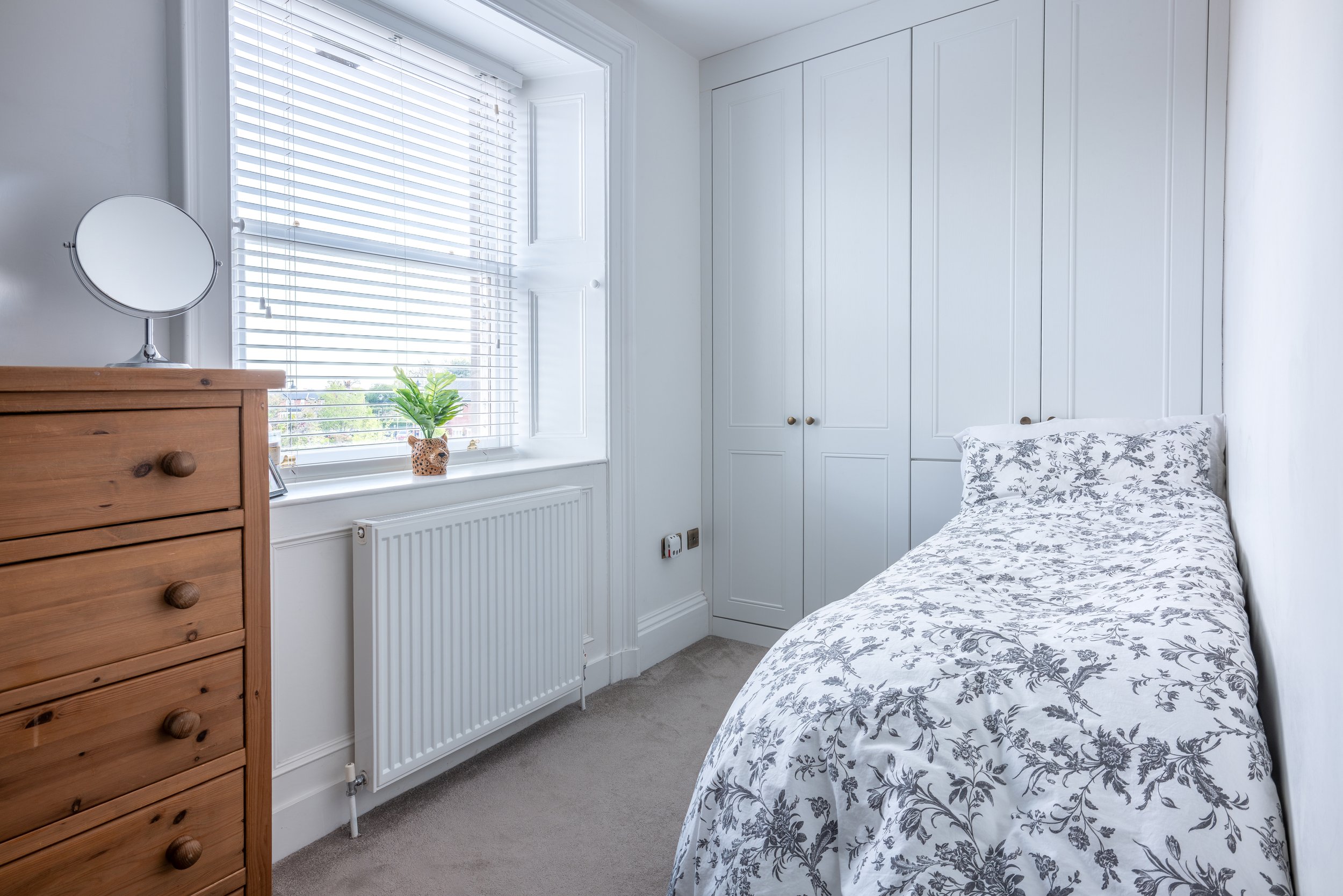 A cozy bedroom with a bed covered in a floral duvet, a white wardrobe, a wooden dresser, a small round mirror on the dresser, a window with white blinds, a small potted plant, and a wall-mounted radiator.
