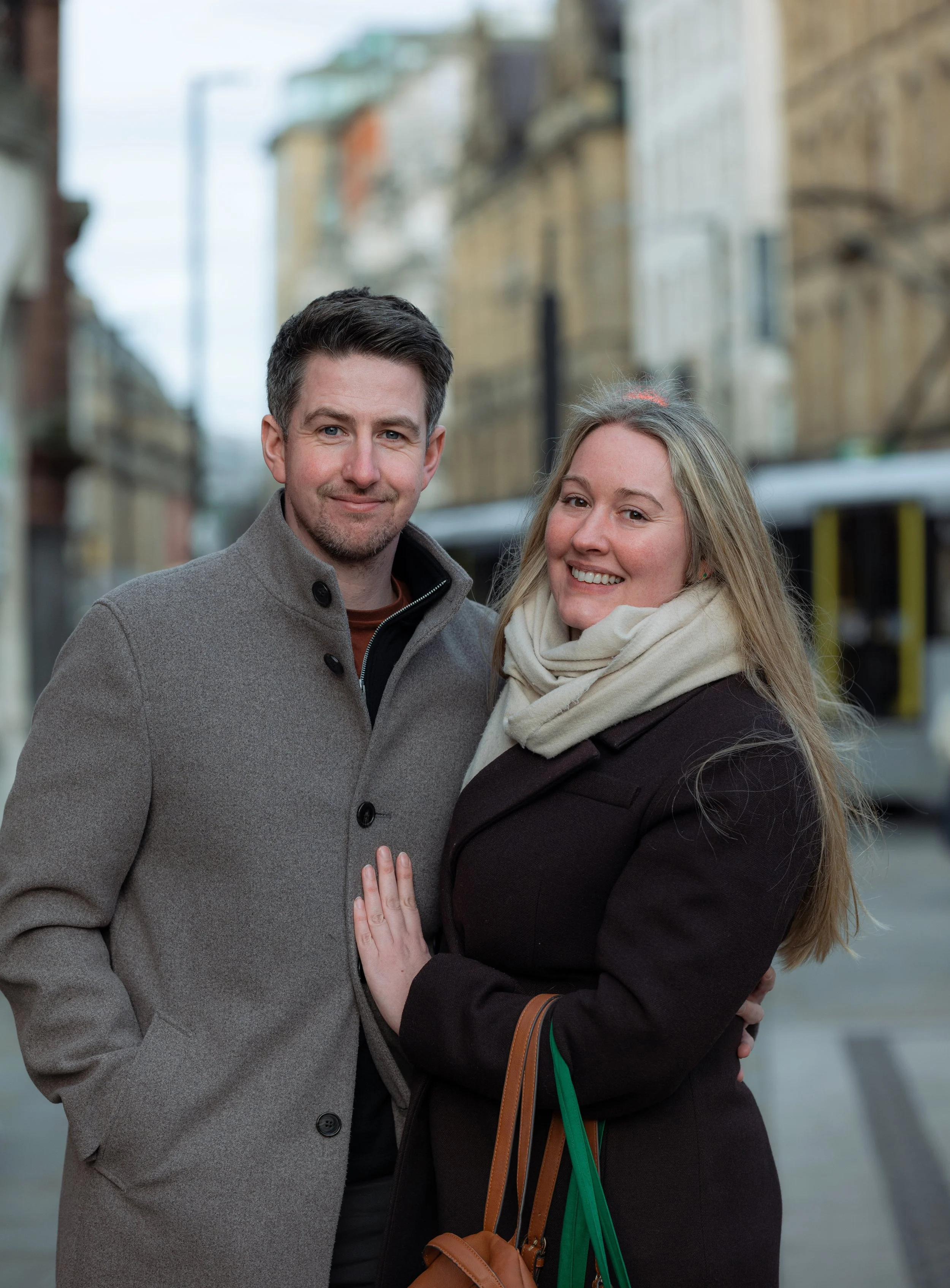 A smiling man and woman standing close together outdoors in an urban setting with buildings and a street in the background. The man is wearing a gray coat, and the woman is wearing a dark coat and a light scarf.