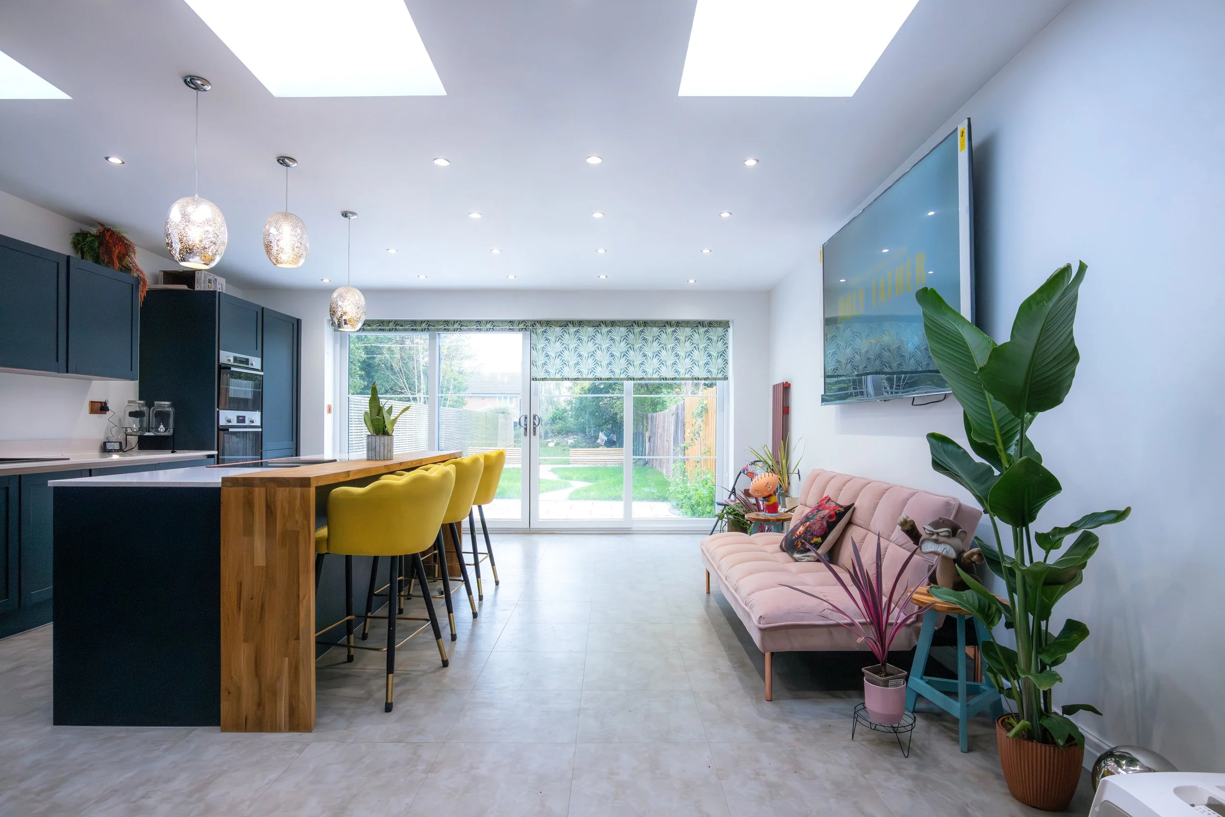 Modern open-plan kitchen and living room with a large wooden island, yellow bar stools, a pink sofa, several potted plants, and a large flat-screen TV on the wall. Sliding glass doors lead to a backyard patio.