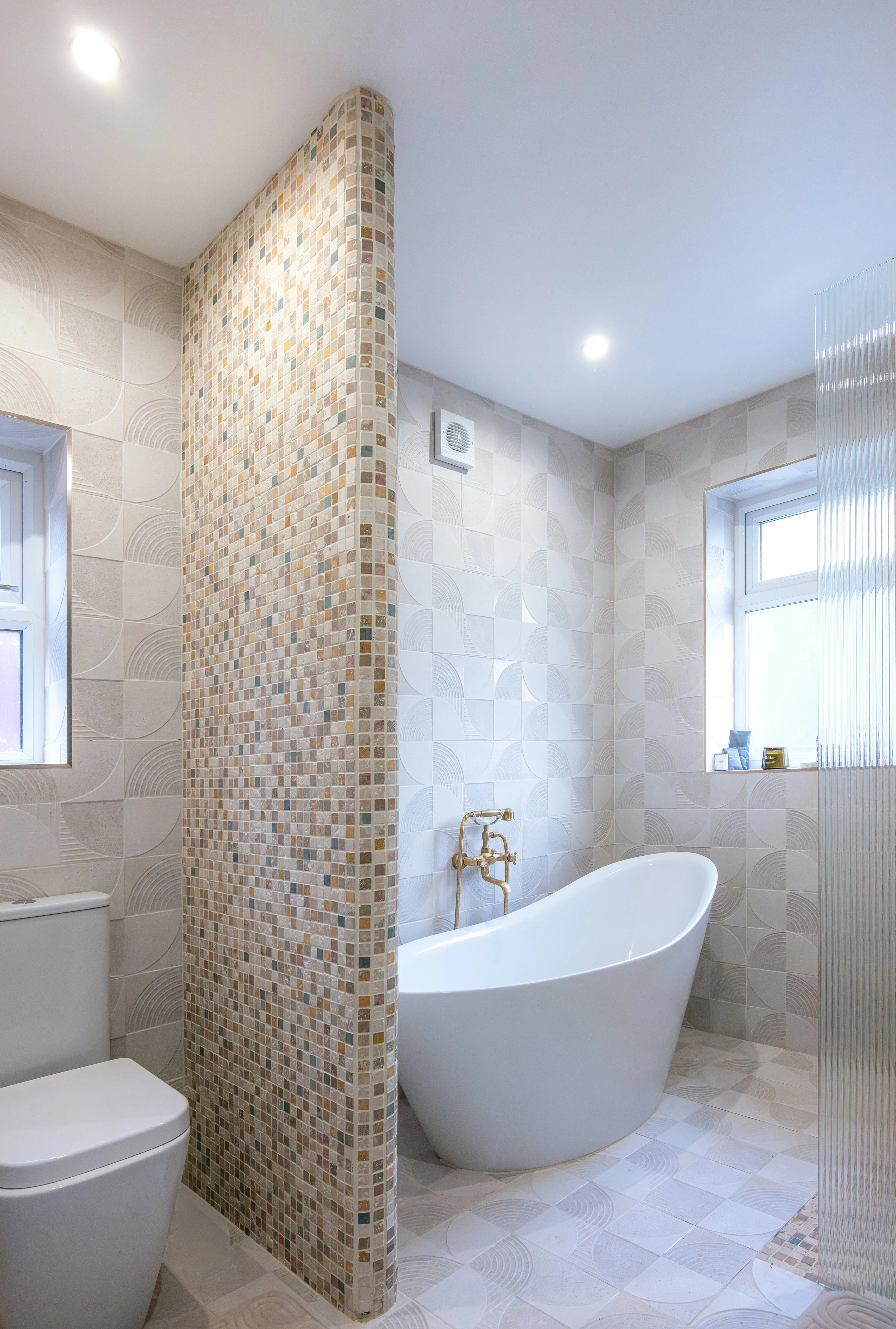 Modern bathroom with a freestanding bathtub, a tiled wall, small window, and a corner partition with mosaic tiles.