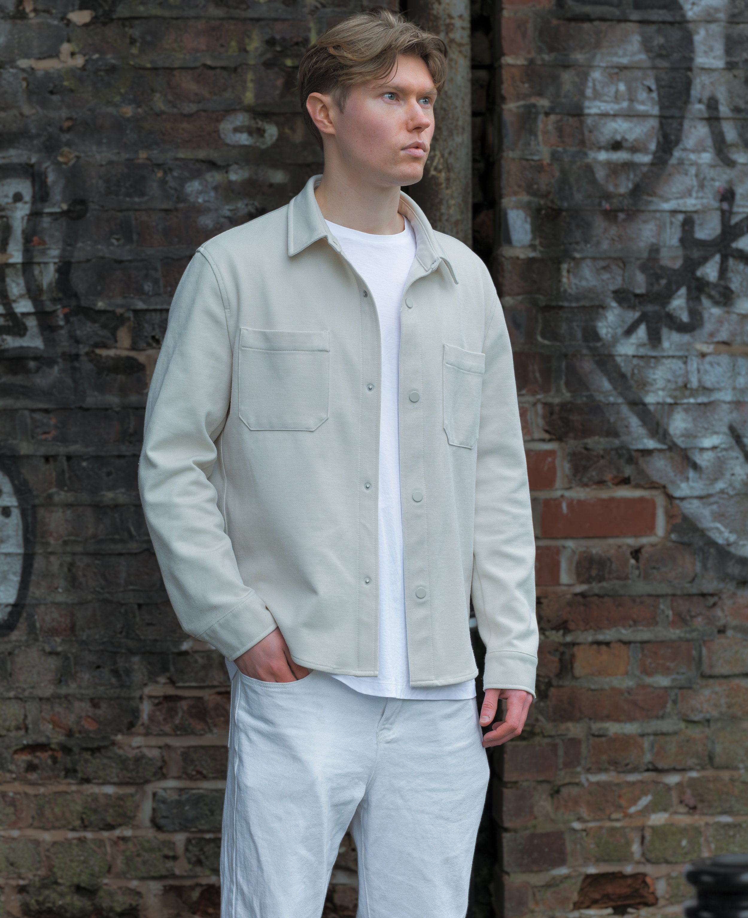 Young man standing outdoors against a graffiti-covered brick wall, wearing a light beige jacket over a white T-shirt and light-colored pants, with his hands in his pockets and looking to the side.