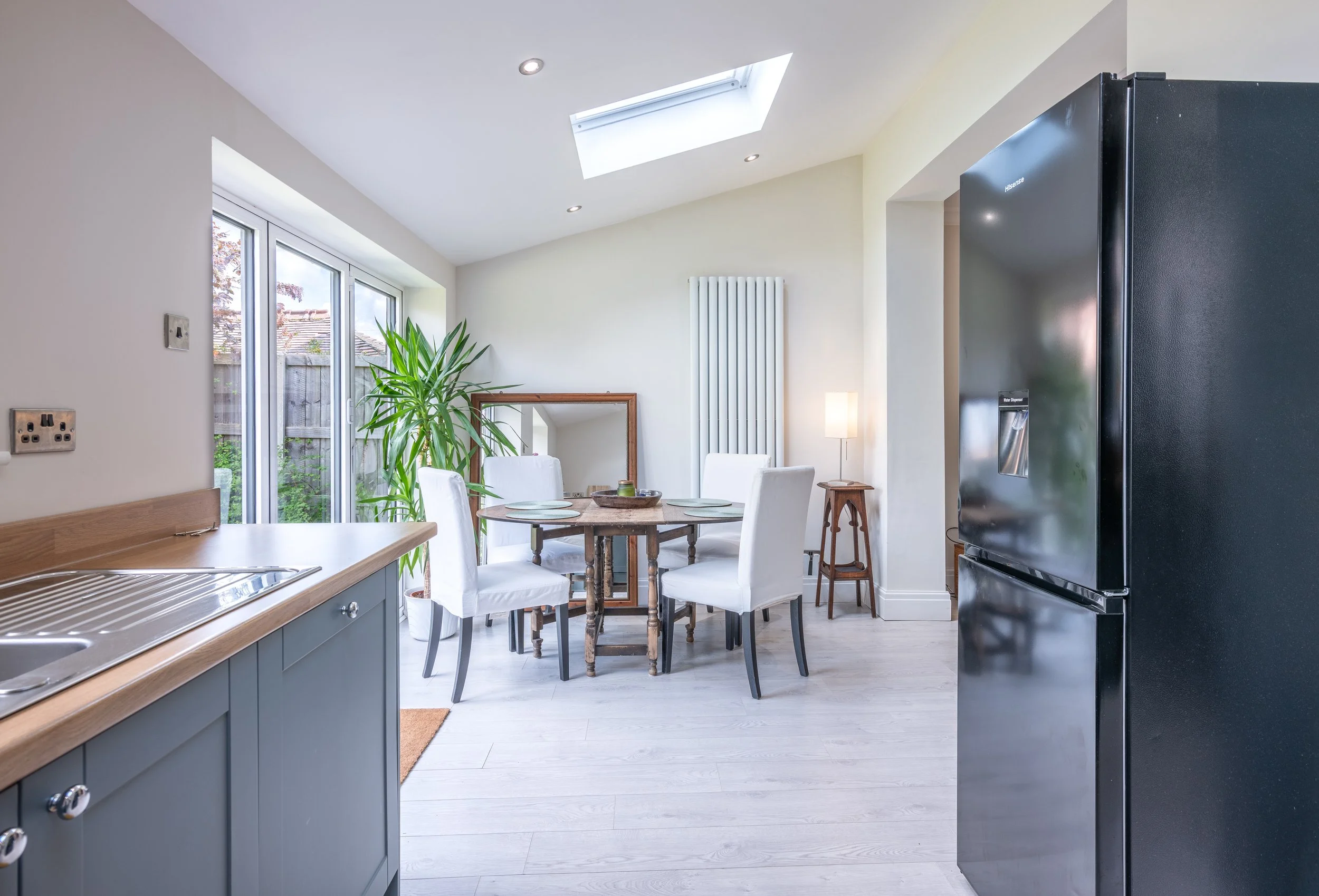 Bright kitchen with round dining table, four white chairs, large window, sliding glass door, indoor plant, skylight, small floor lamp, refrigerator, and wooden flooring.