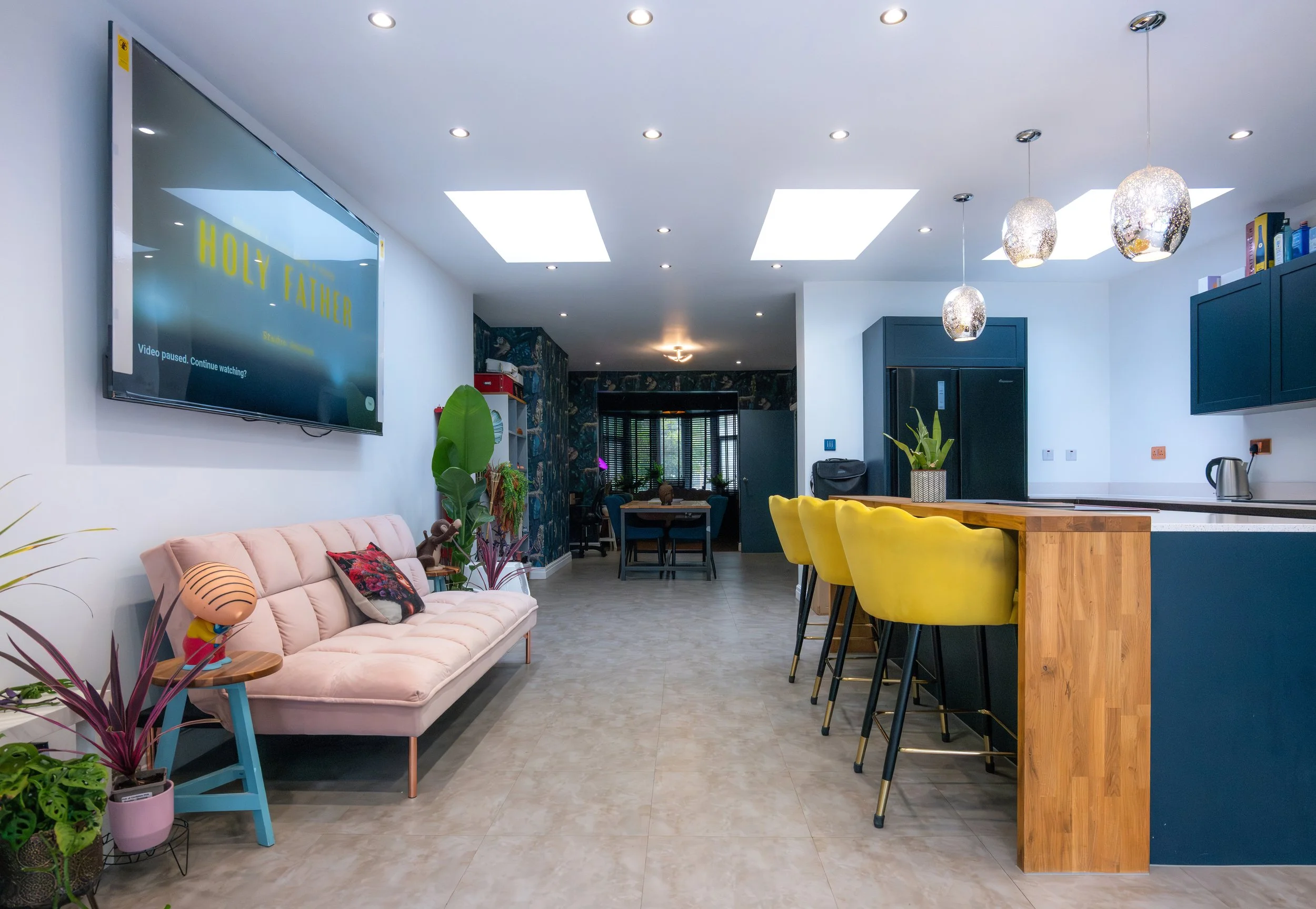 Modern open-concept living room and kitchen with a pink sofa, yellow bar stools, large television, plants, and skylights.