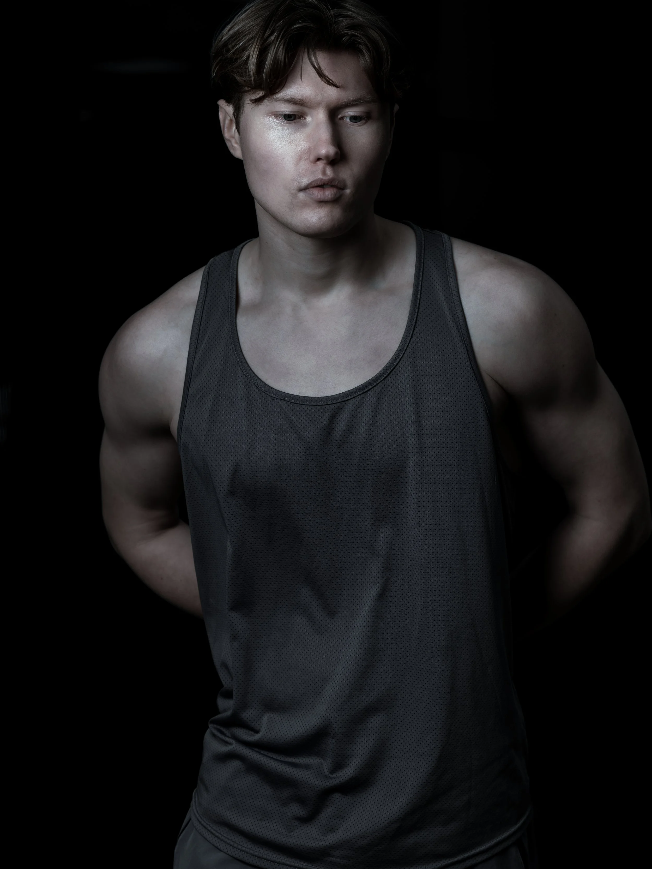 Young man with short brown hair and fair skin, wearing a black athletic tank top, looking down with a neutral expression, against a black background.