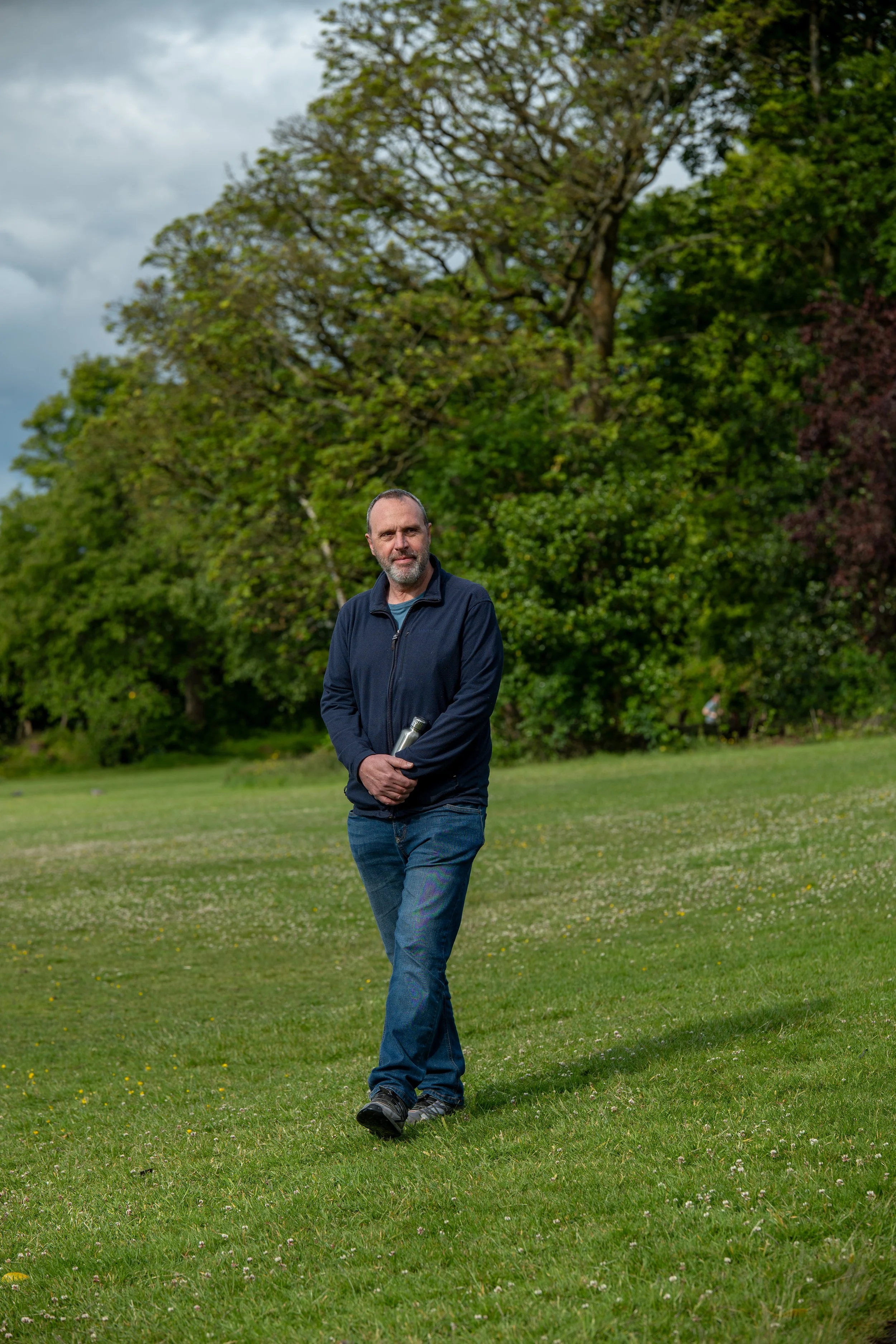 Man walking on grassy field holding a rolled-up paper or magazine, with large trees and cloudy sky in the background.