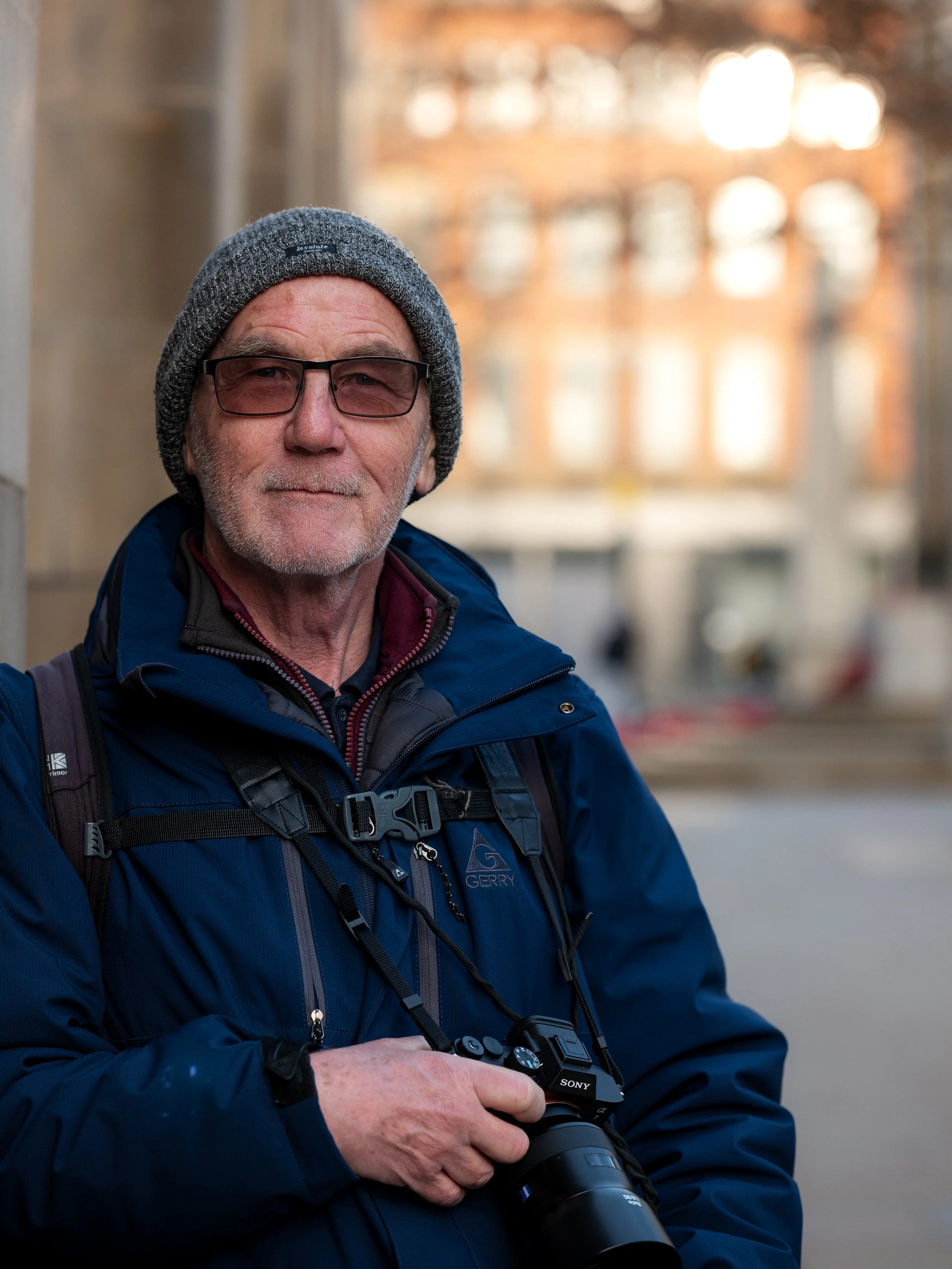An older man with glasses, a gray beanie, and a blue jacket holding a camera outdoors with a blurred city background.