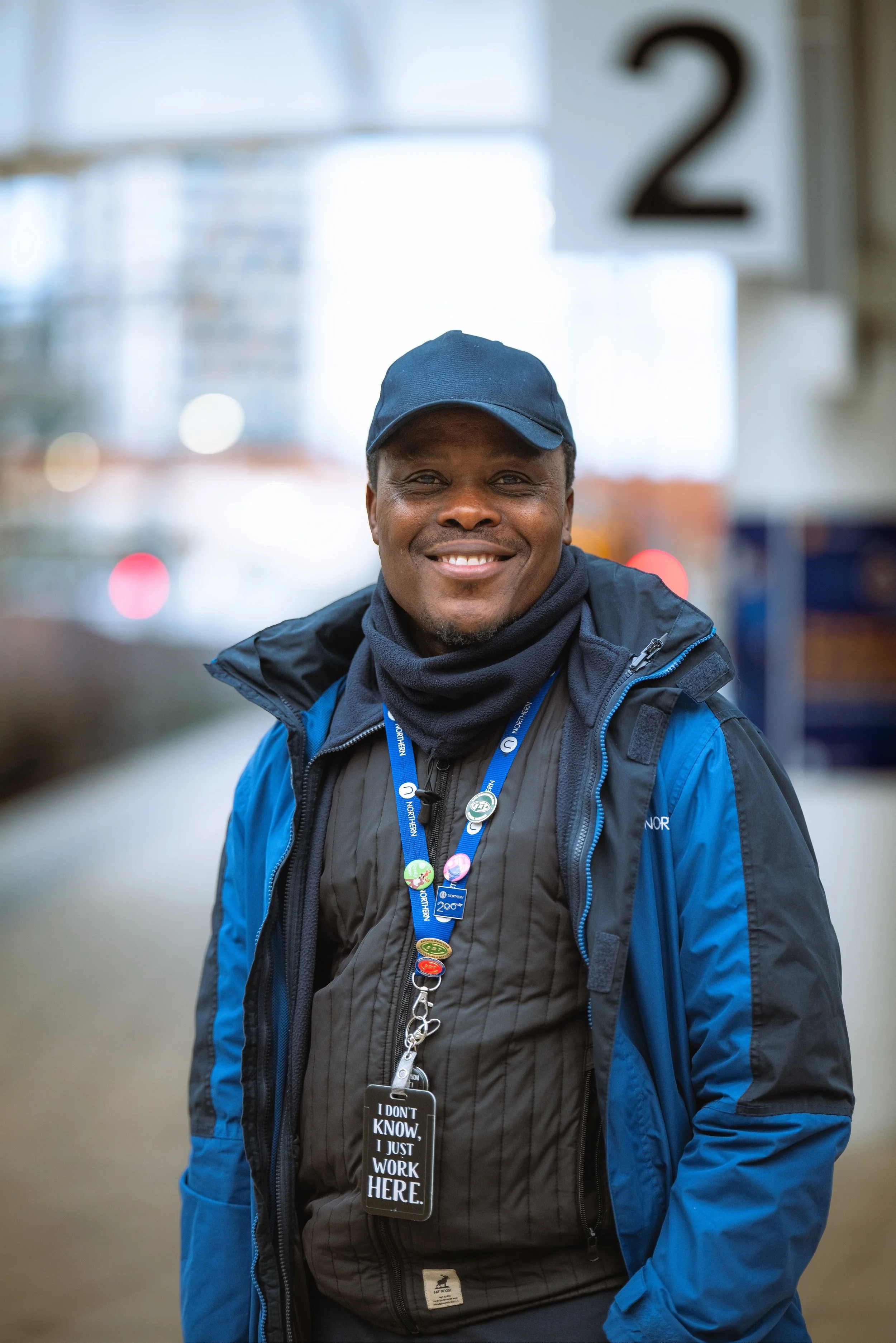 Smiling man wearing a blue jacket, dark scarf, black vest with pins, and a lanyard with a badge that reads "I DON'T KNOW, I JUST WORK HERE." standing outdoors with a blurred background and a sign with the number 2 hanging behind him.