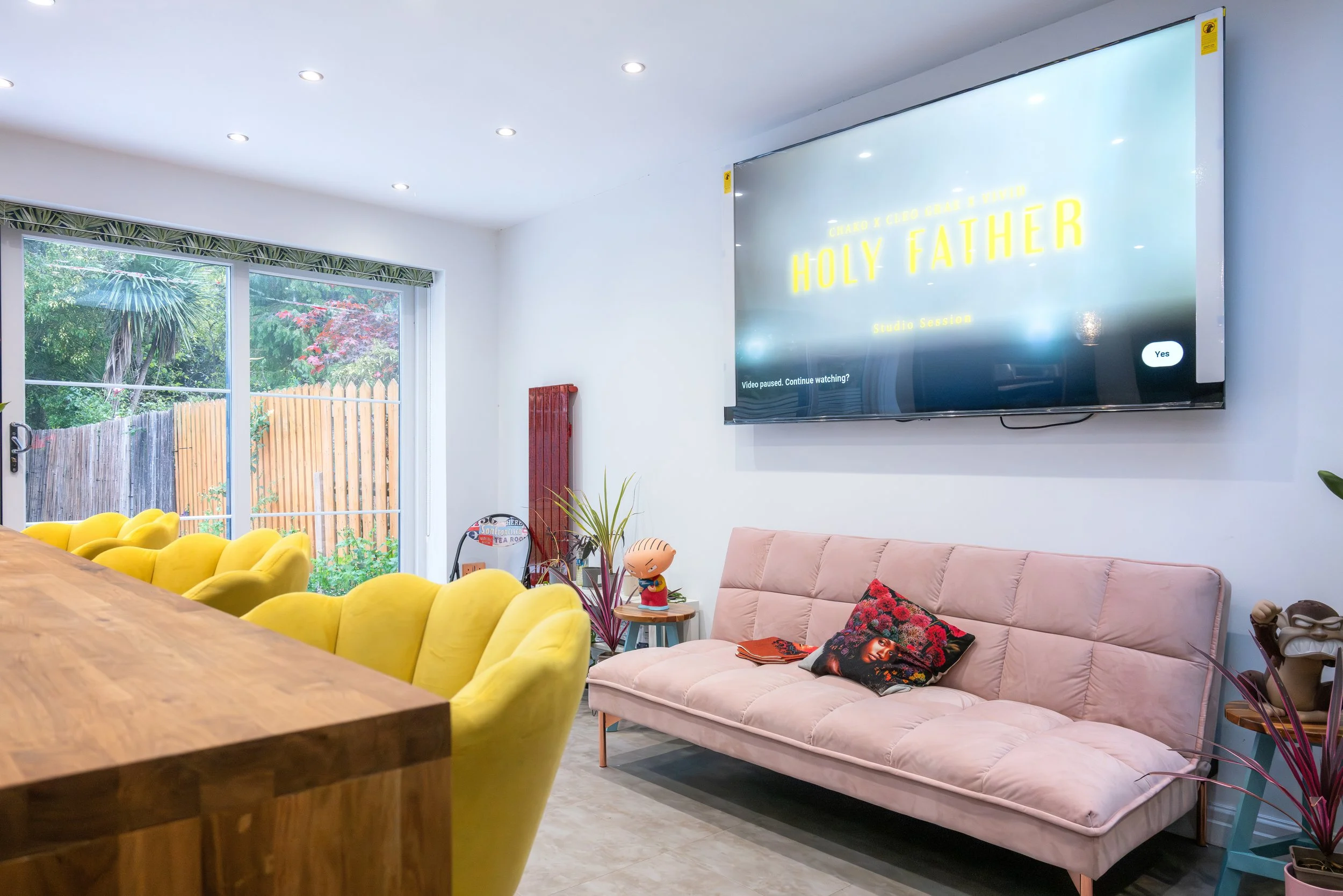 Living room with large glass door, yellow chairs, pink sofa, wall-mounted TV displaying worship service title, and various decorative items including a cartoon character figurine, plants, and artwork.