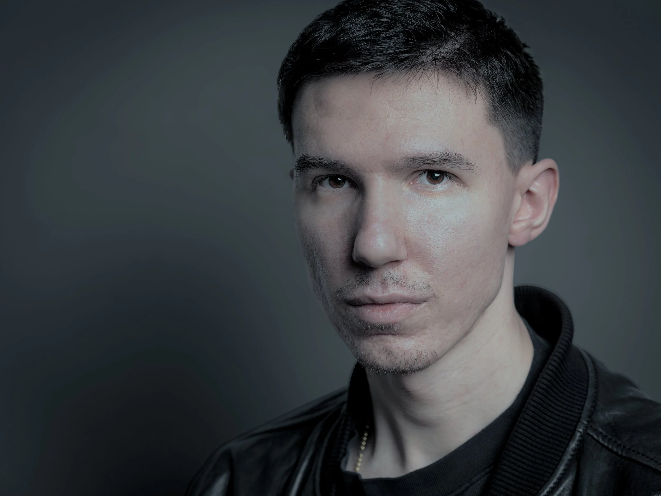 A young man with short dark hair, pale skin, and wearing a black leather jacket looks directly at the camera against a dark gray background.