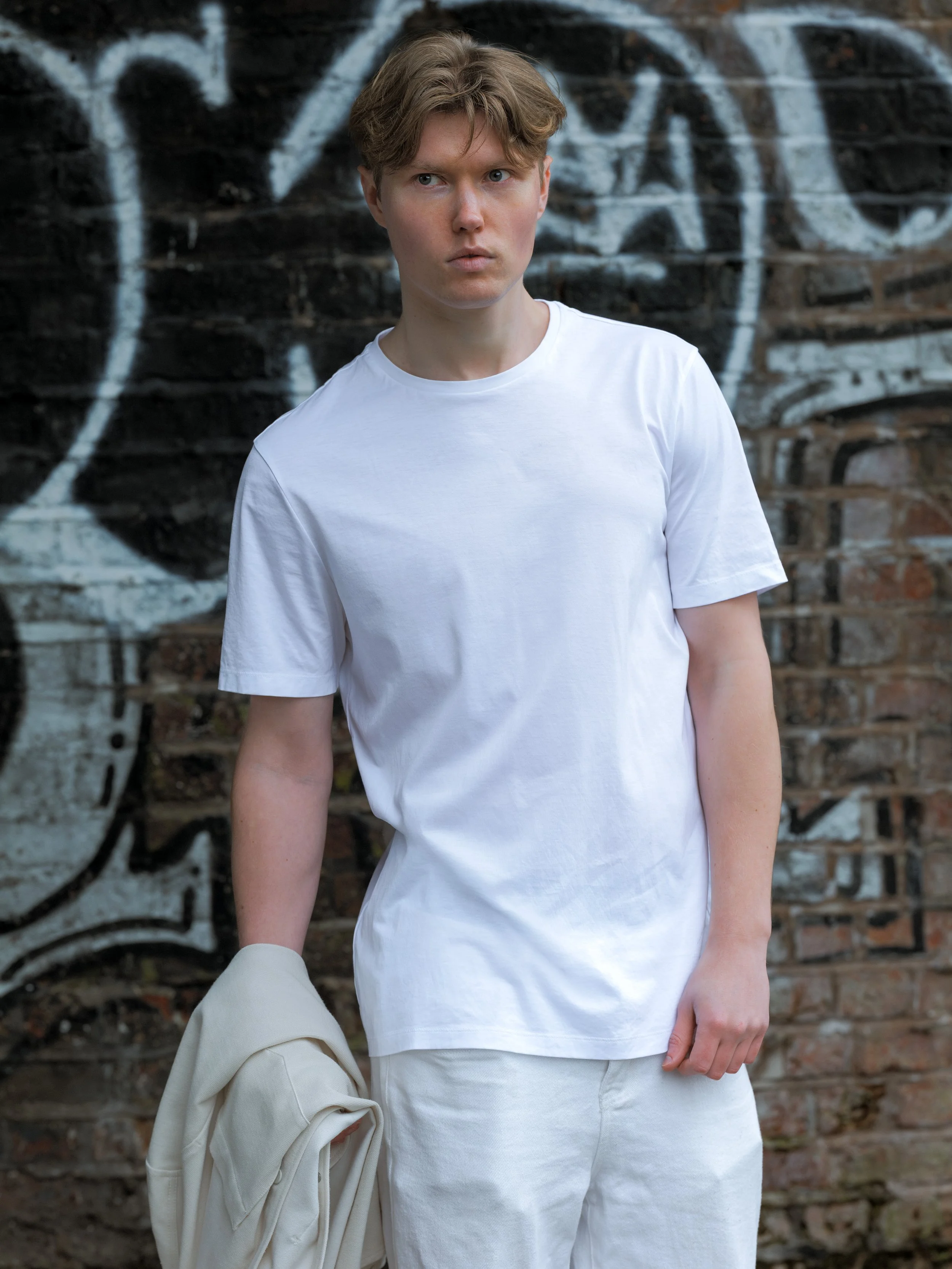 A young man with curly hair and a serious expression, wearing a white t-shirt and light-colored pants, holding a cream-colored jacket, standing in front of a brick wall with graffiti.