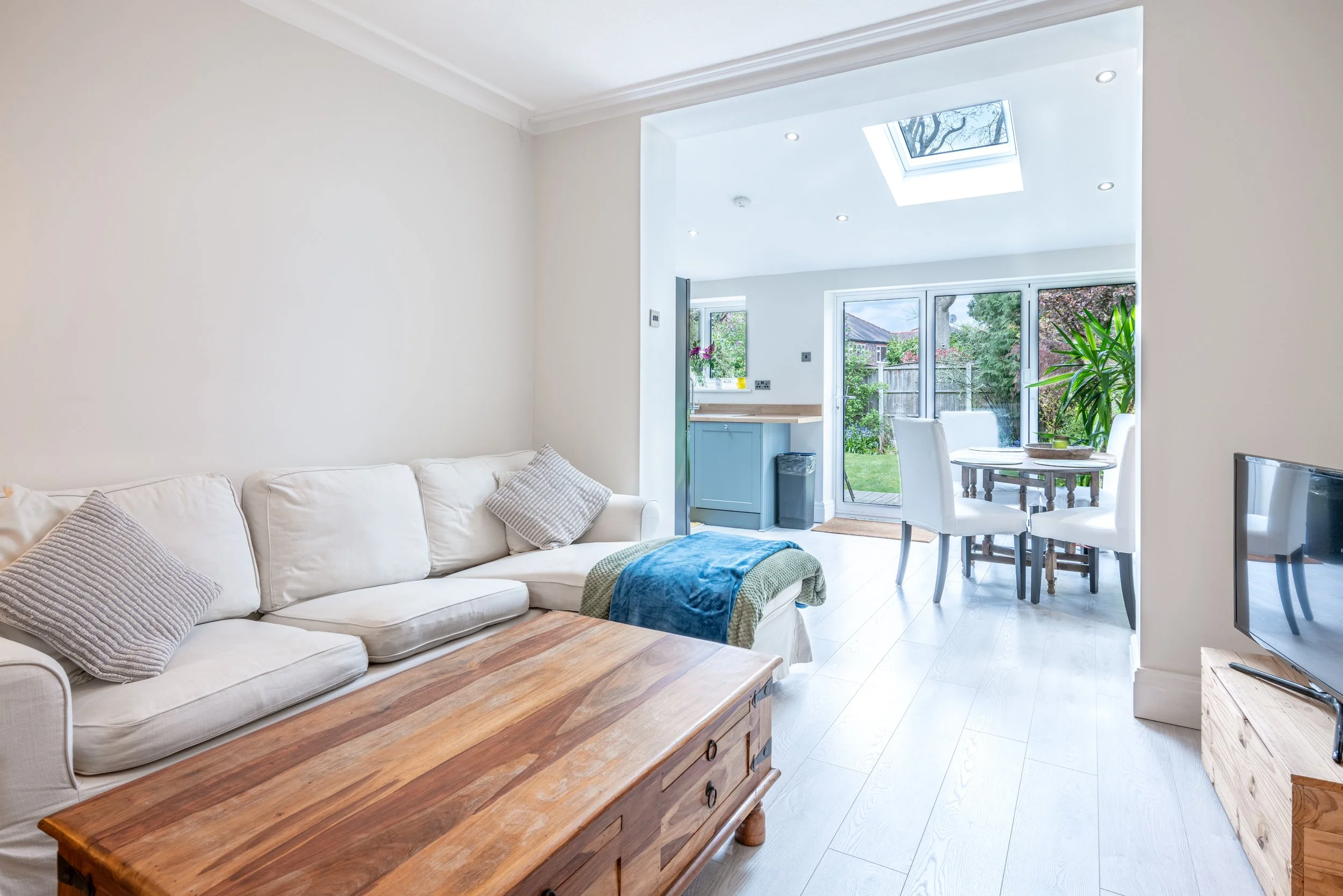 Bright living room with white sofa, wooden coffee table, dining area with white chairs, large window, and garden view.