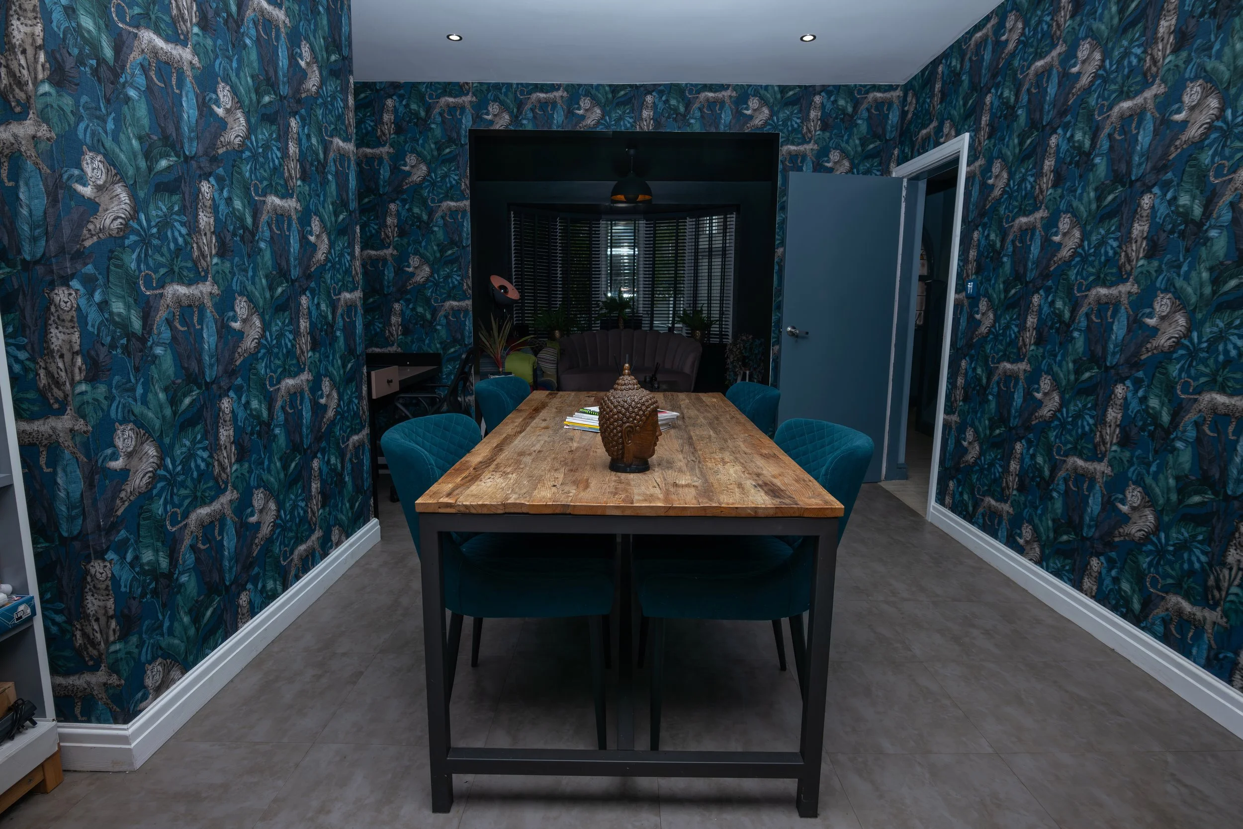 A dining room with a wooden table surrounded by six green upholstered chairs. The walls are covered with jungle-themed wallpaper featuring tigers and leaves. There is a Buddha head sculpture centerpiece on the table, with books and papers nearby. In 