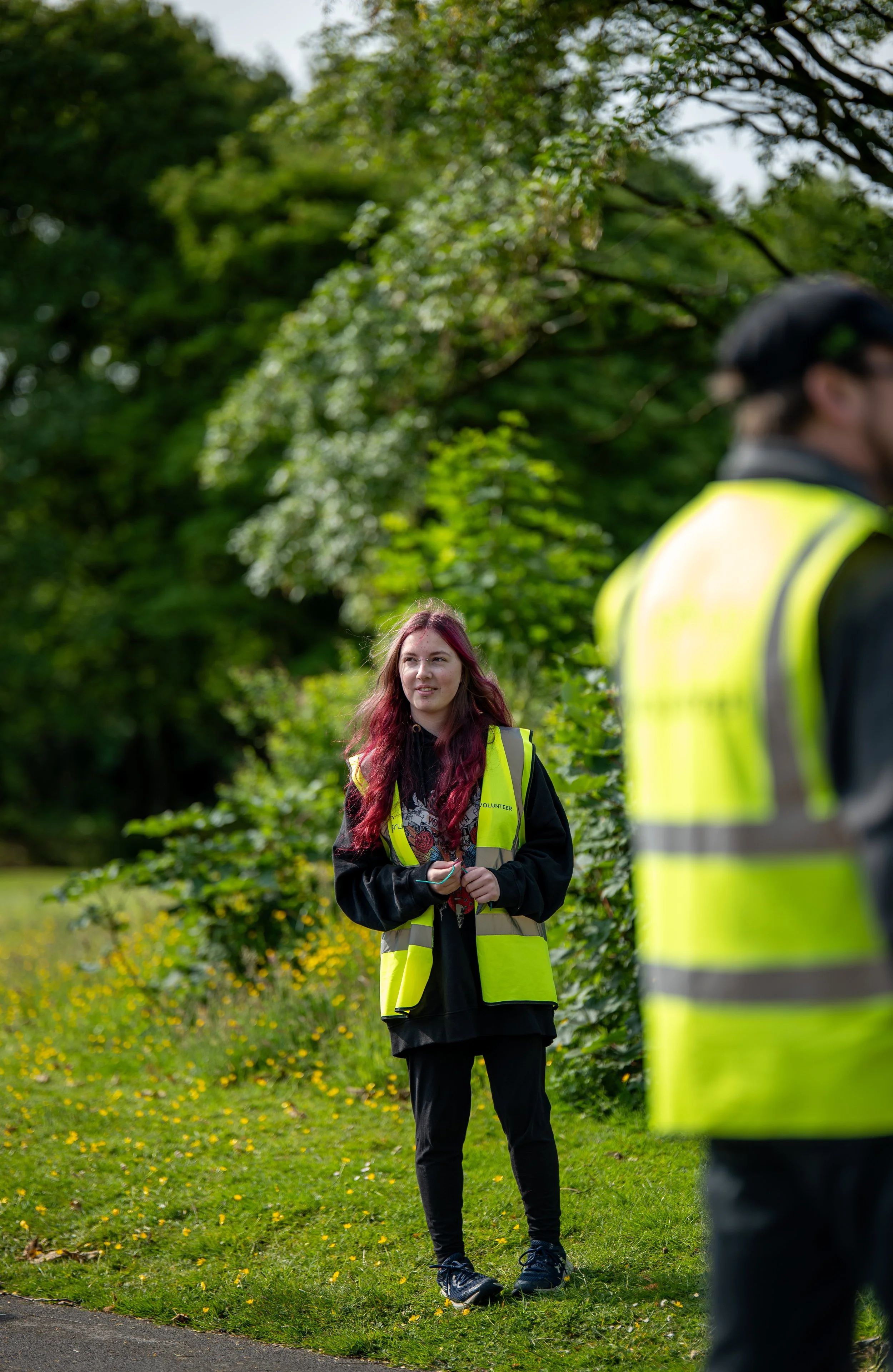 A young woman with pink hair wearing a yellow volunteer vest standing on a grassy area with yellow flowers, speaking to a man in a yellow vest, surrounded by lush green trees.