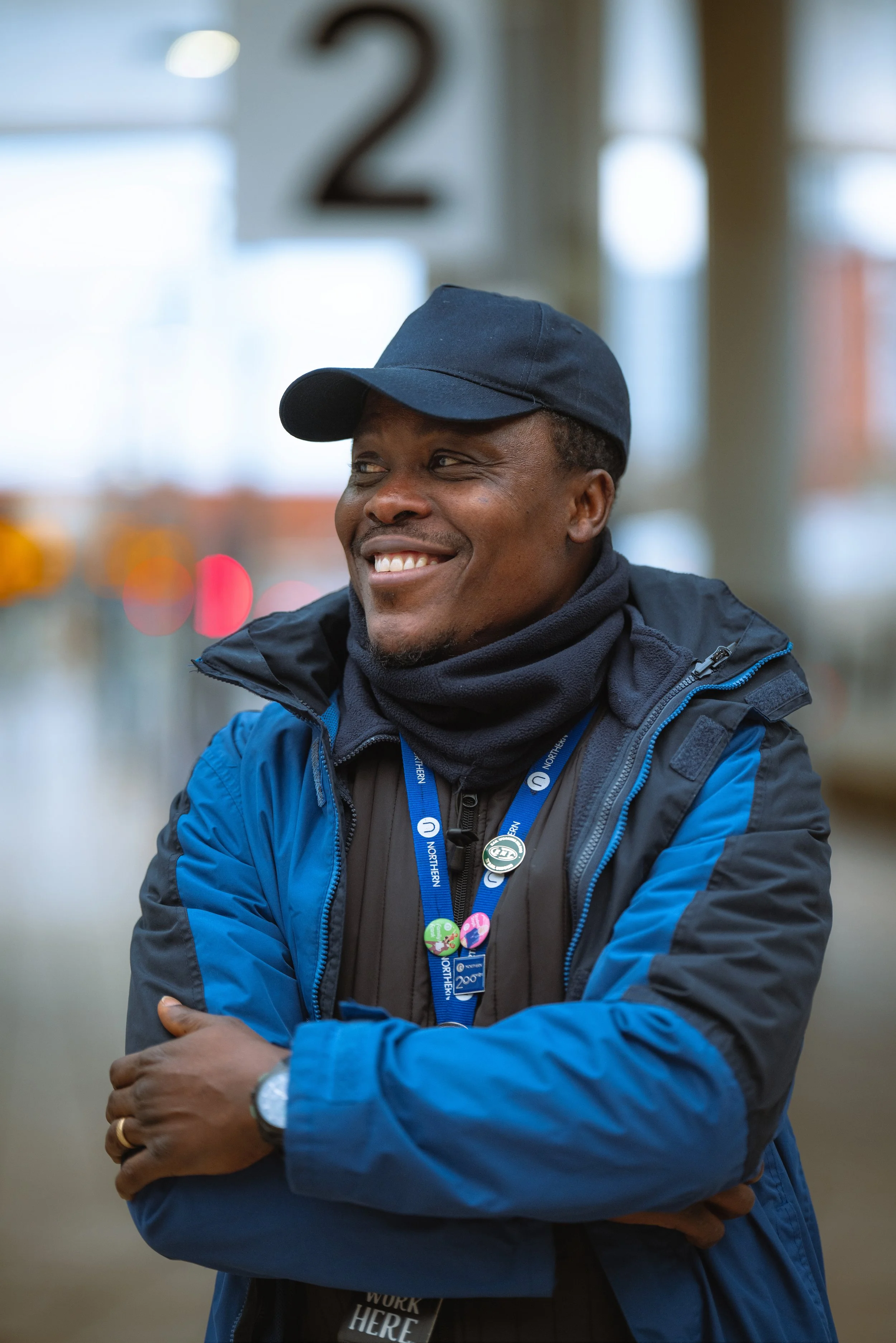 A smiling man in a blue jacket and black baseball cap standing indoors with arms crossed, wearing a navy blue lanyard with badges and a wristwatch.