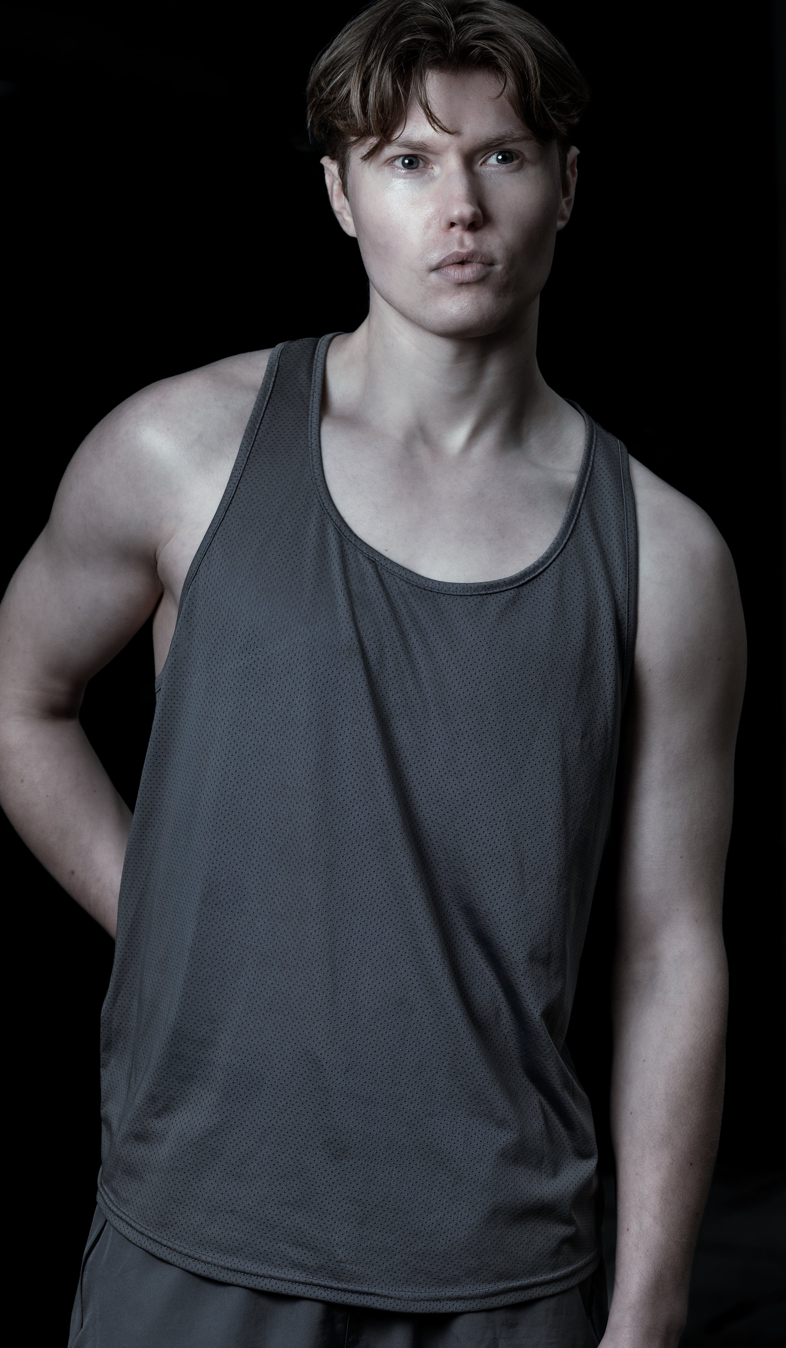 Young man with light skin and short brown hair, wearing a gray athletic tank top, standing against a black background.