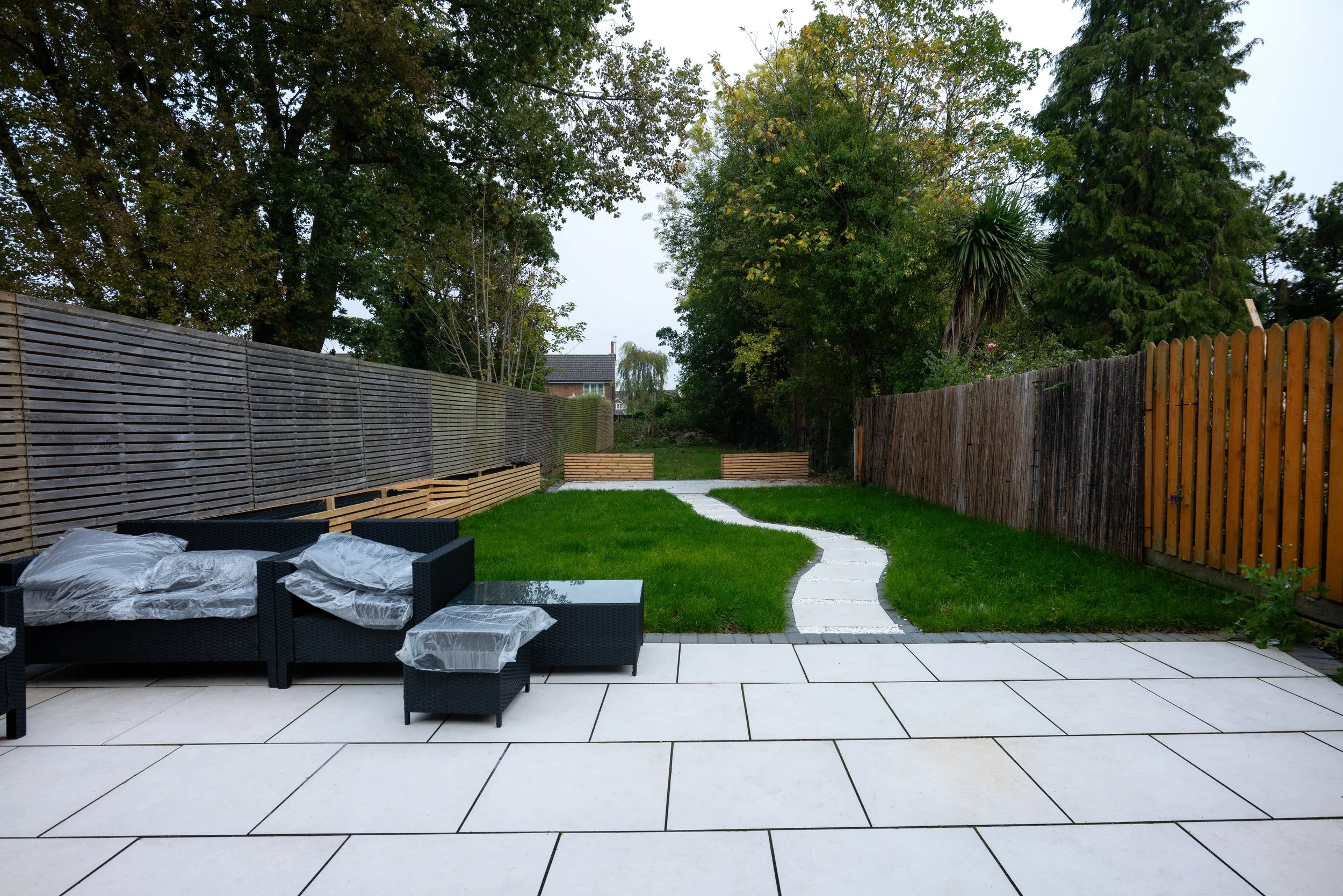Backyard with a tiled patio, black wicker furniture covered with plastic, a winding stone pathway, grass lawn, and a fence on both sides, with trees and a cloudy sky.