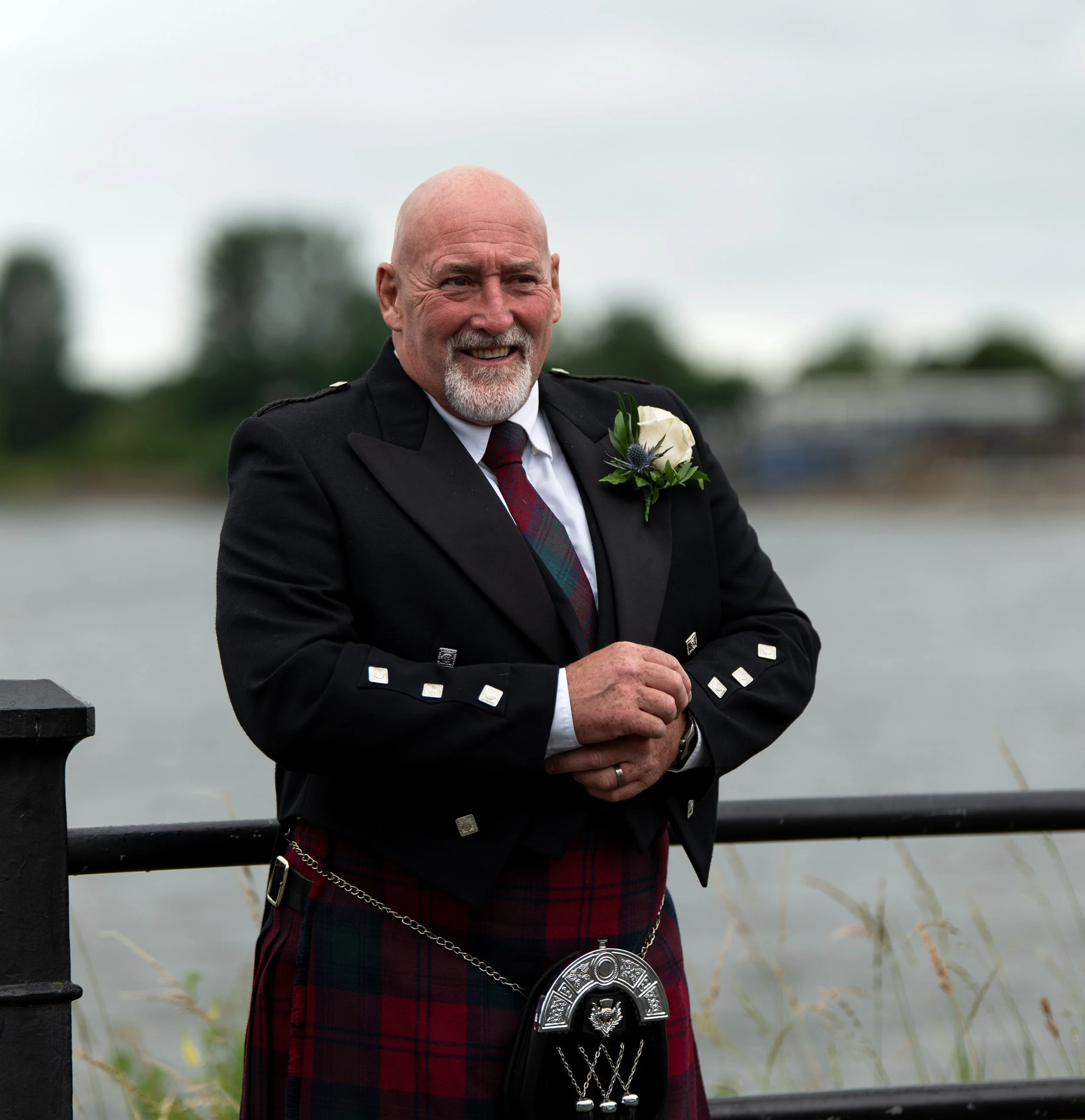 A man wearing a traditional Scottish kilt and black jacket, with a white flower boutonniere, stands outdoors near a black metal railing, smiling, with a calm water body and trees in the background.