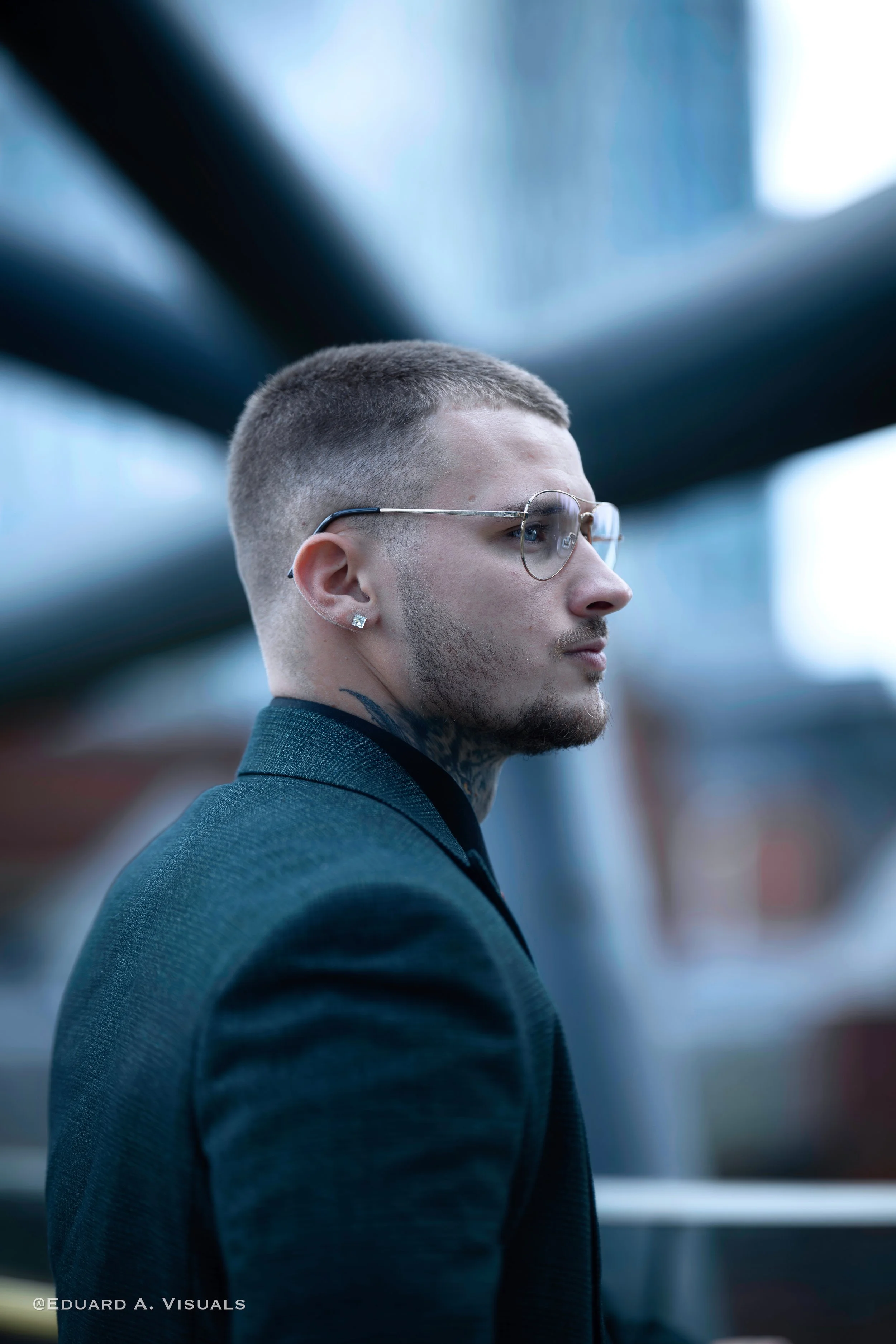 Profile of a young man with short hair, wearing glasses, earrings, a tattoo on his neck, and a suit in an urban setting with blurred background.