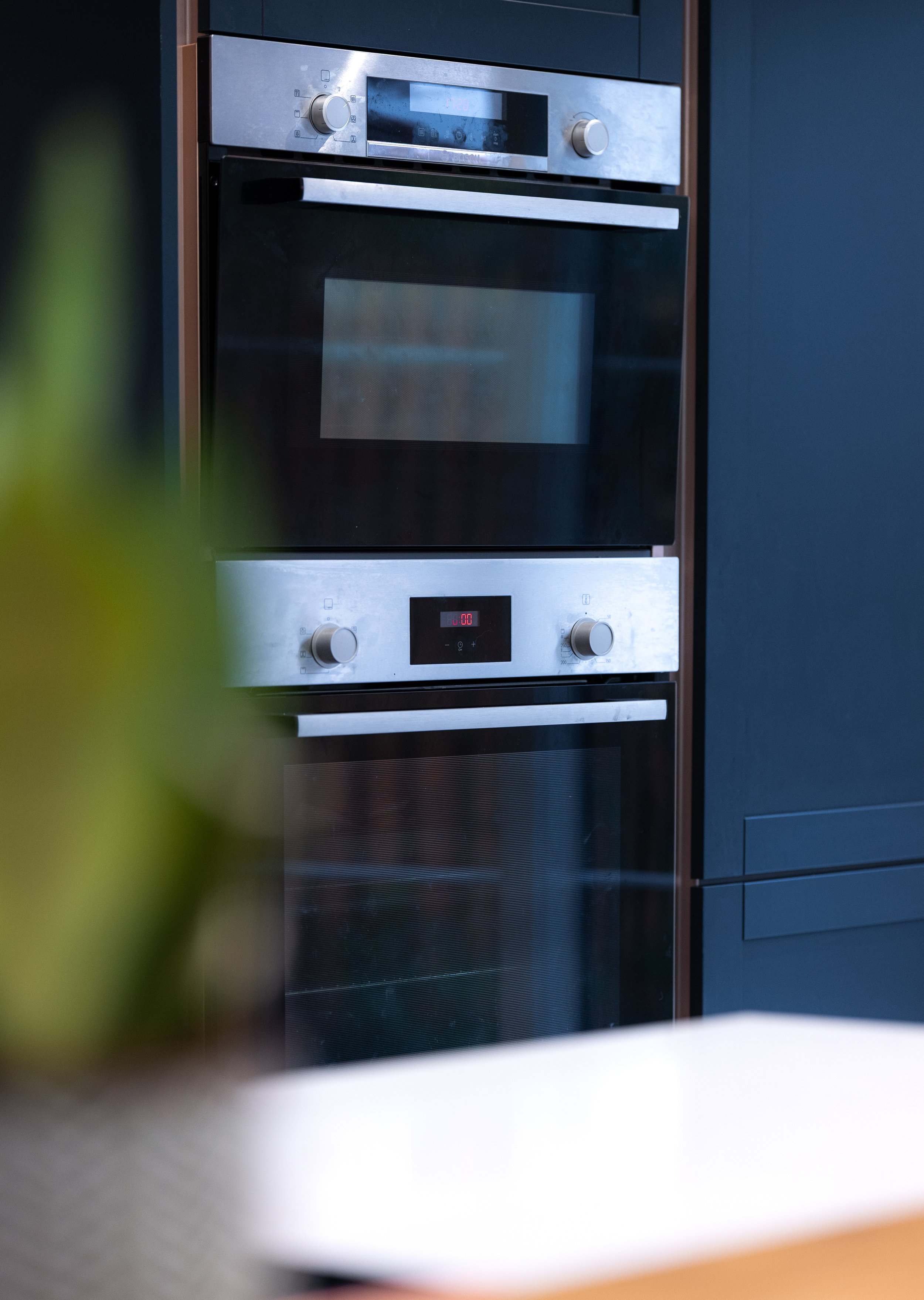 Modern stainless steel and black kitchen oven with control knobs and digital display, partially obscured by a blurred green plant and white surface in the foreground.