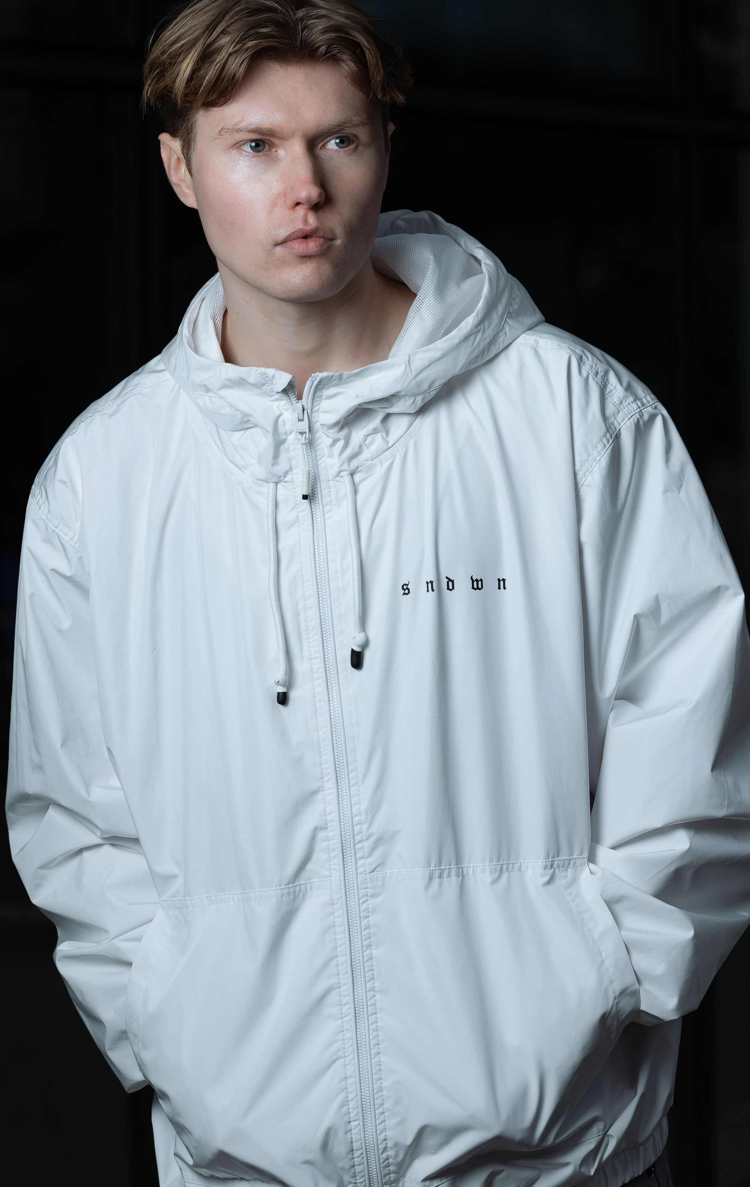 Young man with light skin and brown hair wearing a white windbreaker jacket with the word 'snow' on it, standing in front of a dark background.