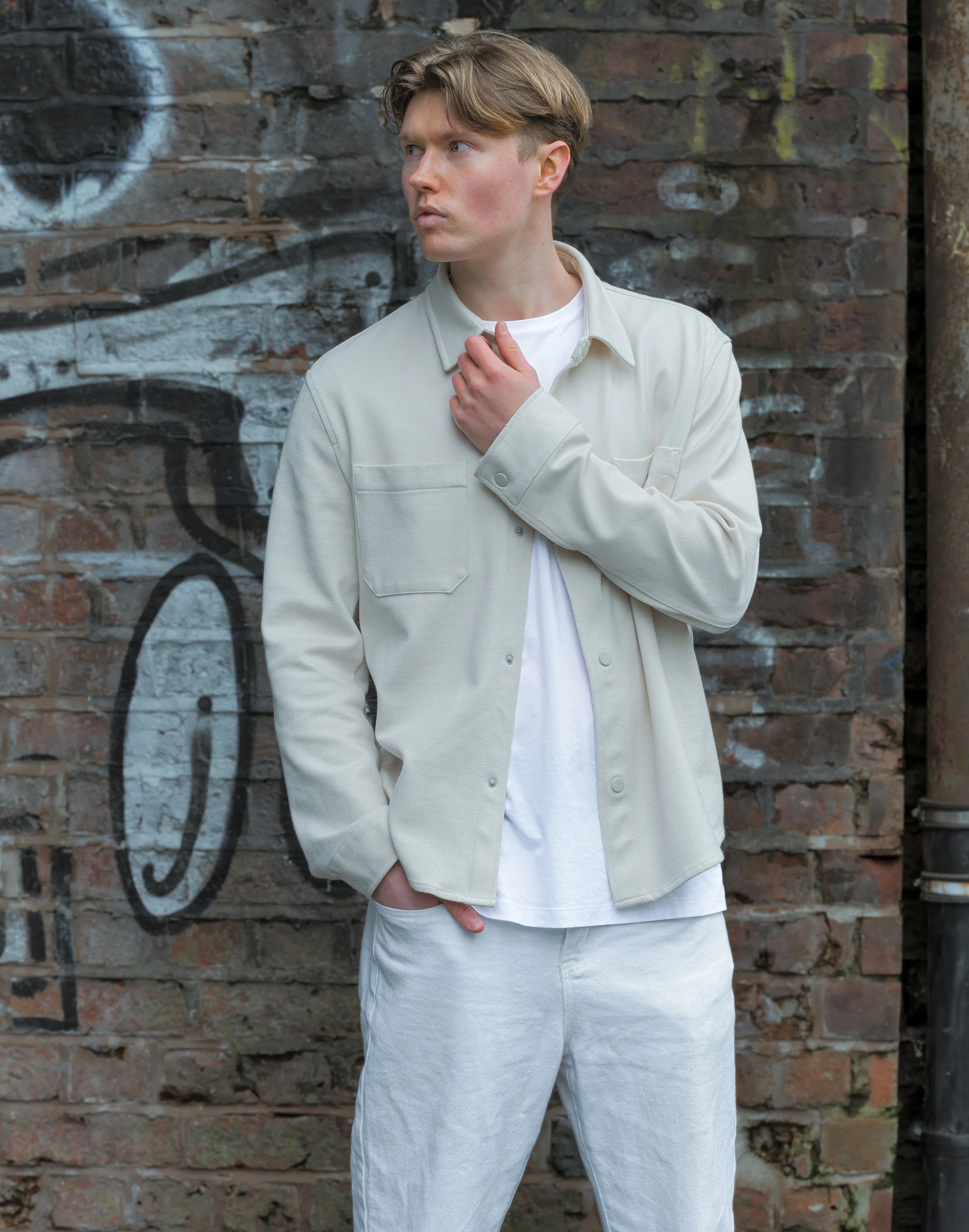 Young man in light-colored jacket and pants standing against a graffiti-covered brick wall.