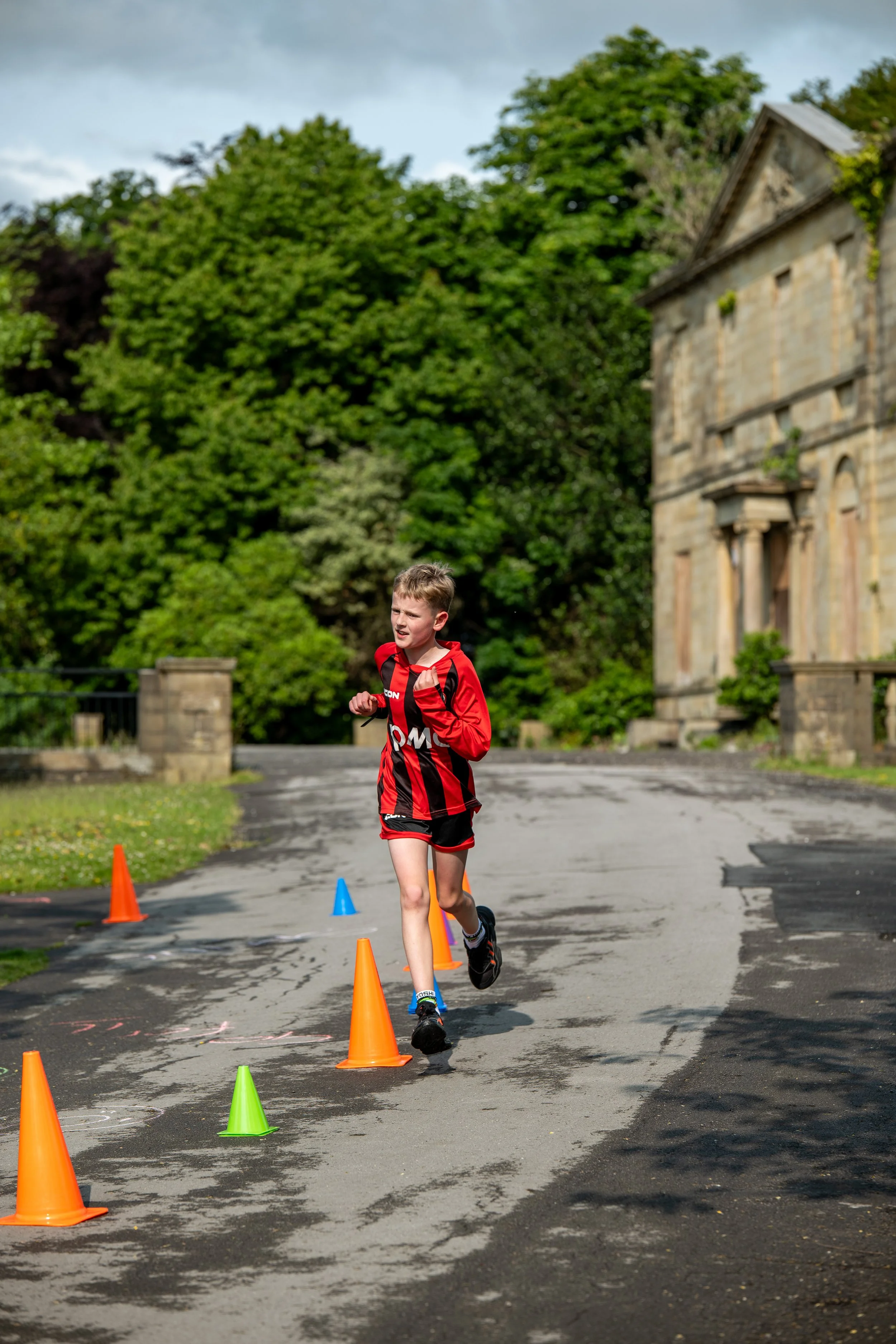 A young boy running on a paved path with colorful cones set up for an outdoor activity, with greenery and an old building in the background.
